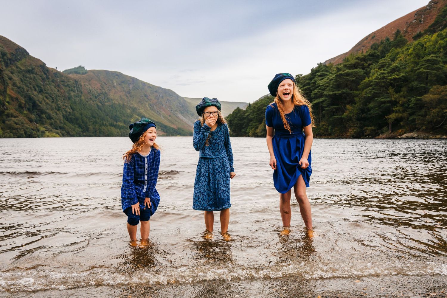 Three people stand ankle-deep in shallow water at a scenic mountain lake on a cloudy day.Family photogrAPHER DUBLIN