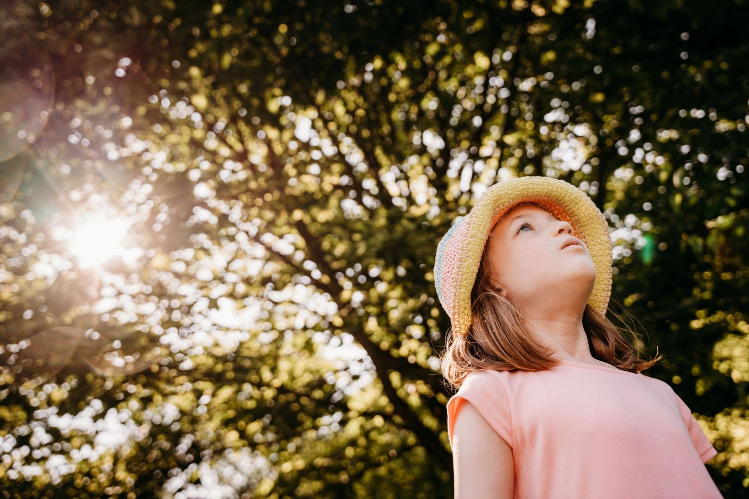 Person in pink top wearing a straw hat gazes up at sunlight streaming through leafy trees in a peaceful outdoor scene. Family PHOTOGRAPHER D