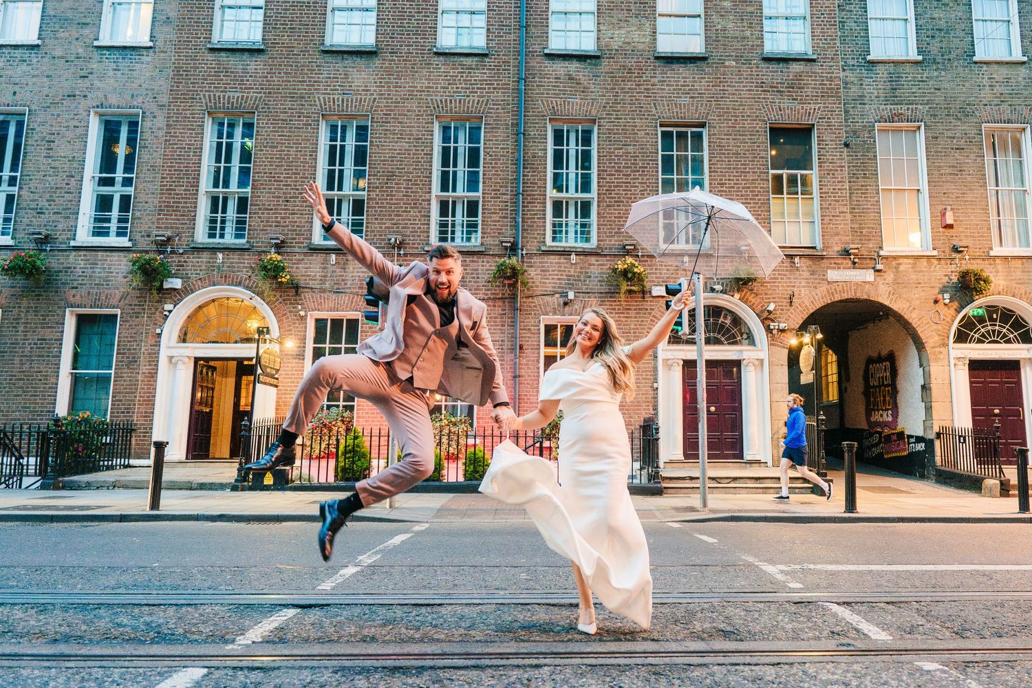 Three people jump joyfully in front of historic Georgian buildings on a Dublin street with brick facade and arched doorways. Wedding PHOTOGR