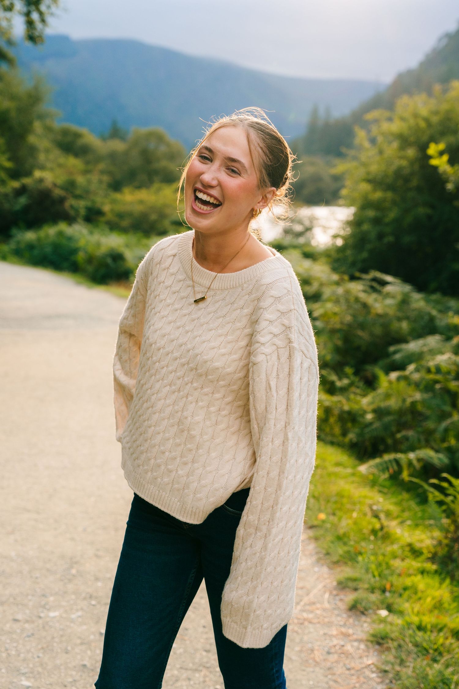 A person in a cozy cream cable knit sweater laughs joyfully on a mountain path surrounded by lush greenery. PHOTOGRAPHER Dublin