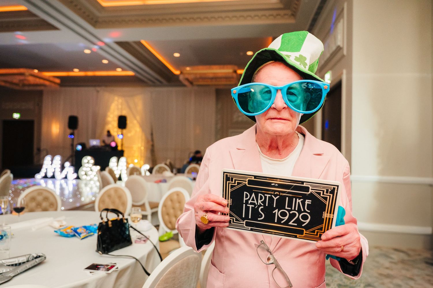 A party guest wearing oversized turquoise sunglasses and green party hat holds up a decorative party sign at a reception.