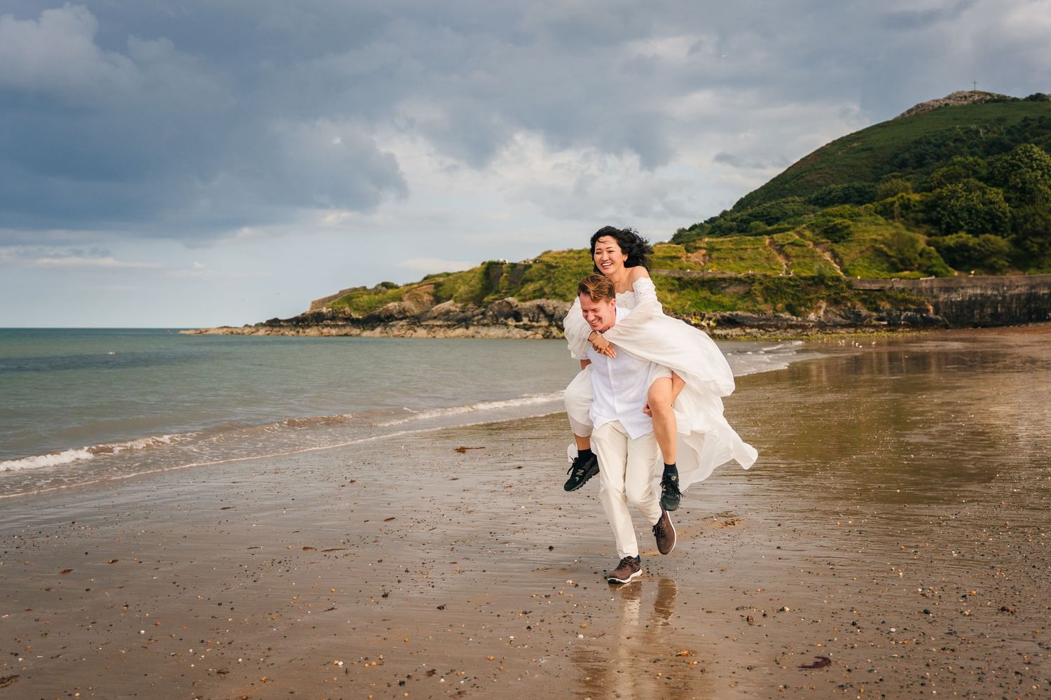 A couple enjoying a romantic moment on a scenic beach with green hills and moody clouds in the background. PHOTOGRAPHER Dublin