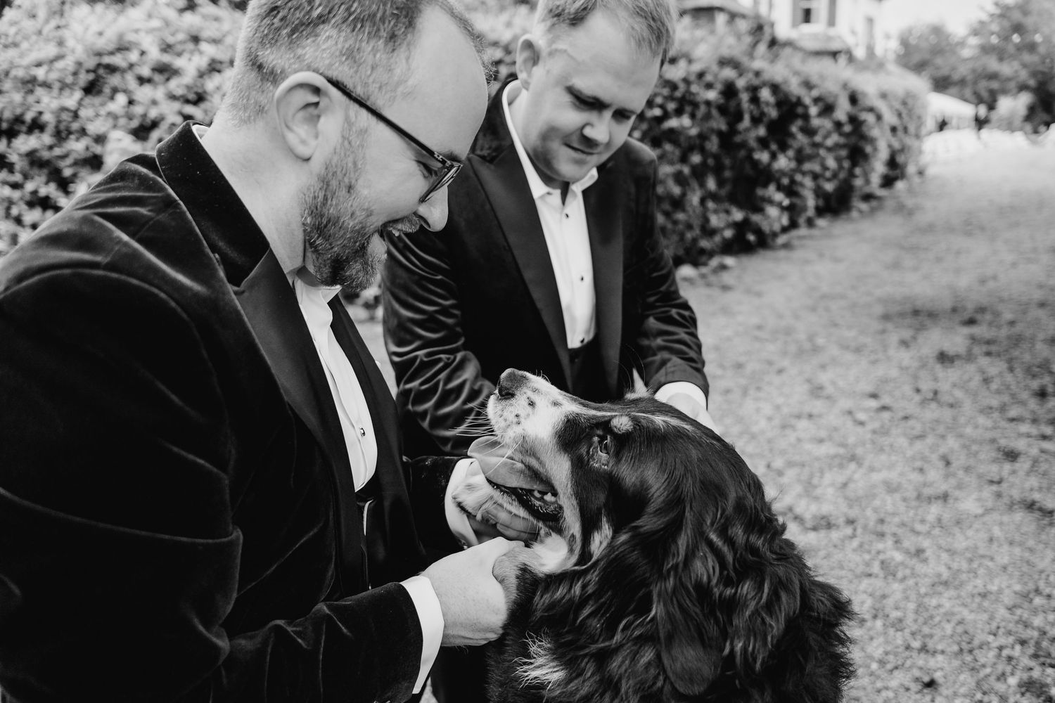 Two people in formal attire petting a black and white dog outdoors. LGBTQ Ashley Park Wedding Photographer