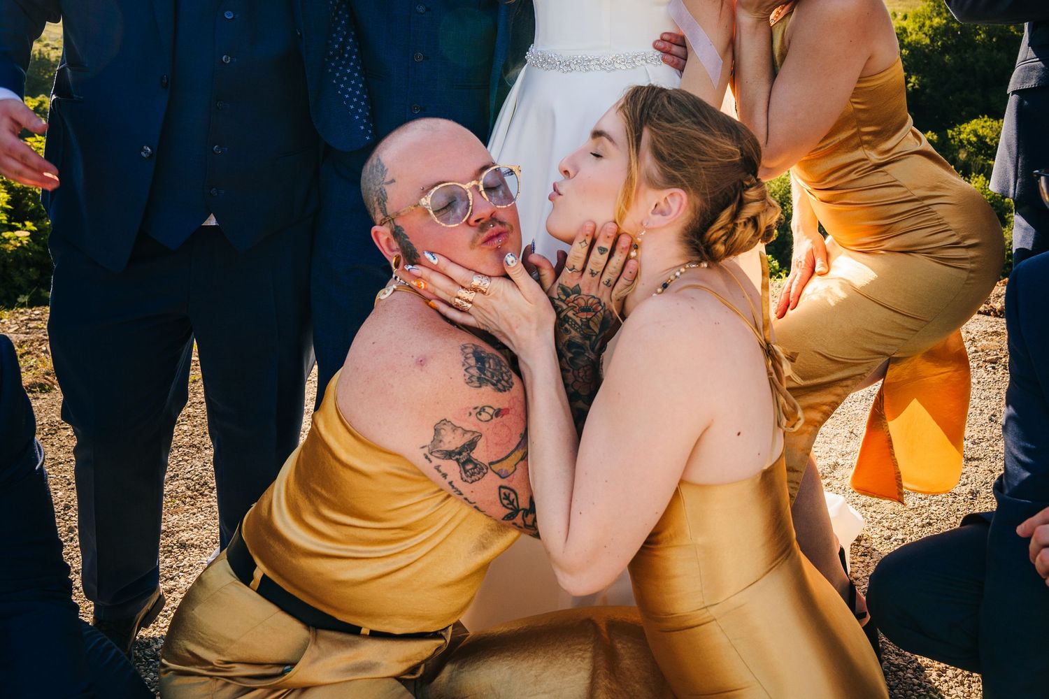 Two bridesmaids in gold dresses share a playful moment at a wedding celebration outdoors.