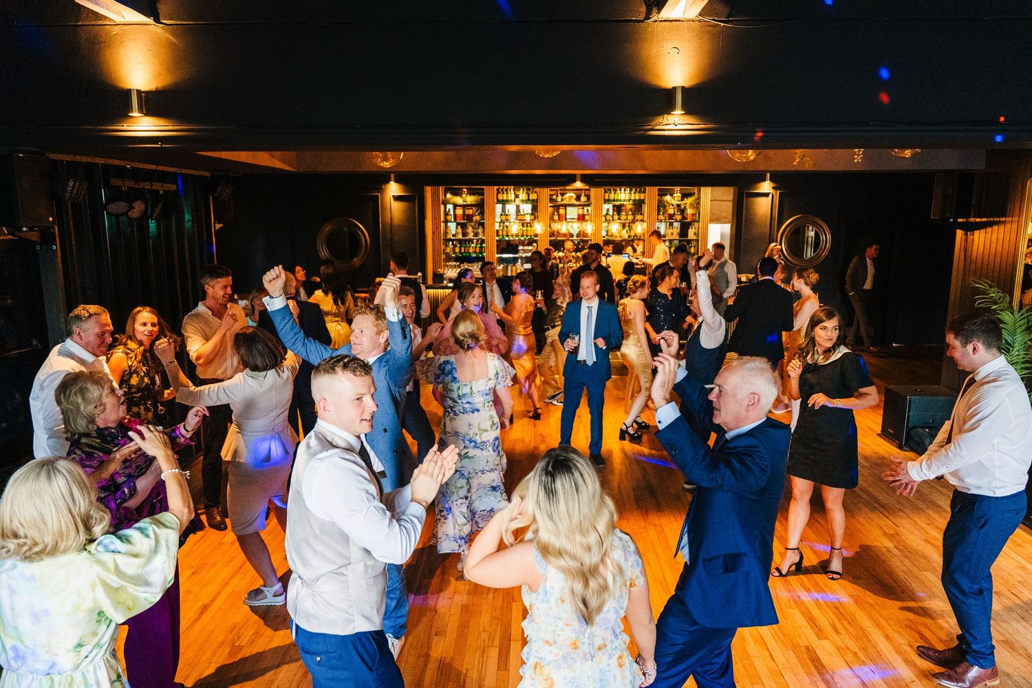 Wedding party guests dance together on a wooden dance floor under warm lighting at an evening reception celebration.