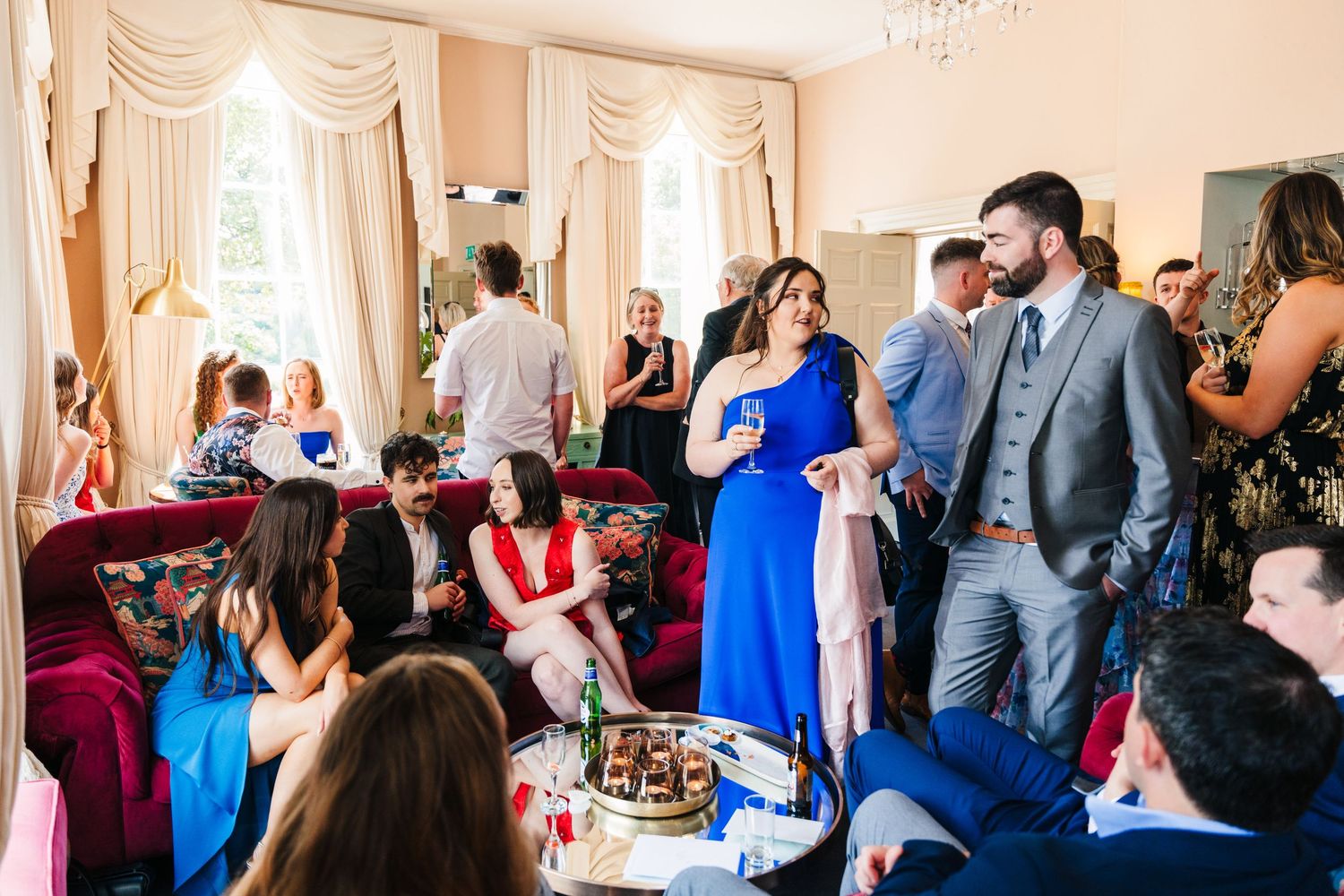 Guests mingle at an elegant celebration in a room with cream curtains and red velvet chairs during a social gathering.Millhouse slane weddin