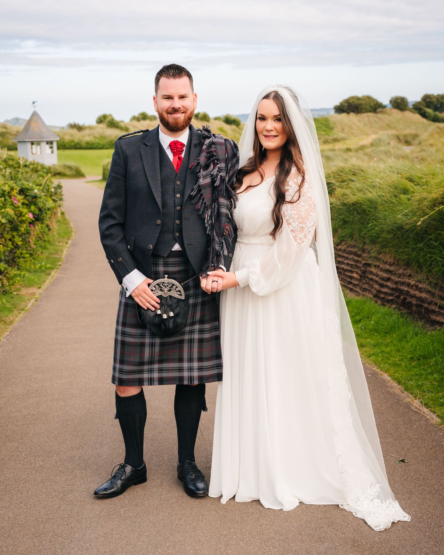 A couple poses on a pathway, the groom in traditional Scottish kilt and the bride in an elegant white wedding gown. Portmarnock Hotel Weddin