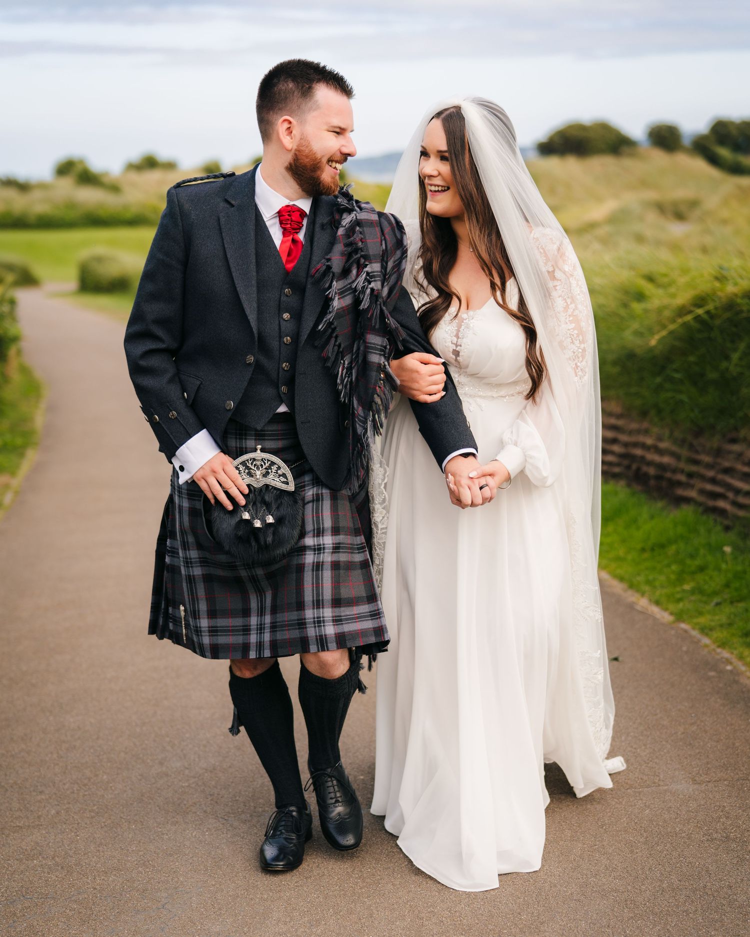 A pair in traditional Scottish wedding attire walking together on a path with rolling green hills in the background. Wedding PHOTOGRAPHER Du