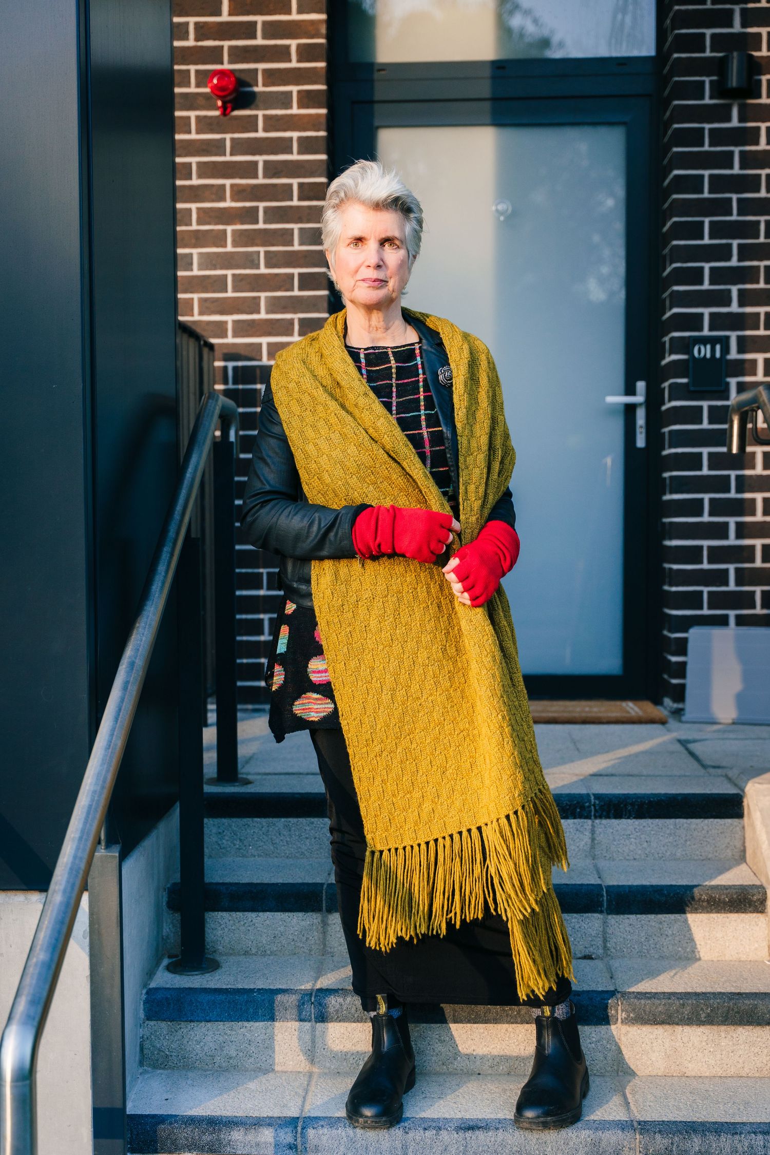 Person wearing a mustard yellow shawl and red gloves sitting on concrete steps by a brick building. professional headshots dublin
