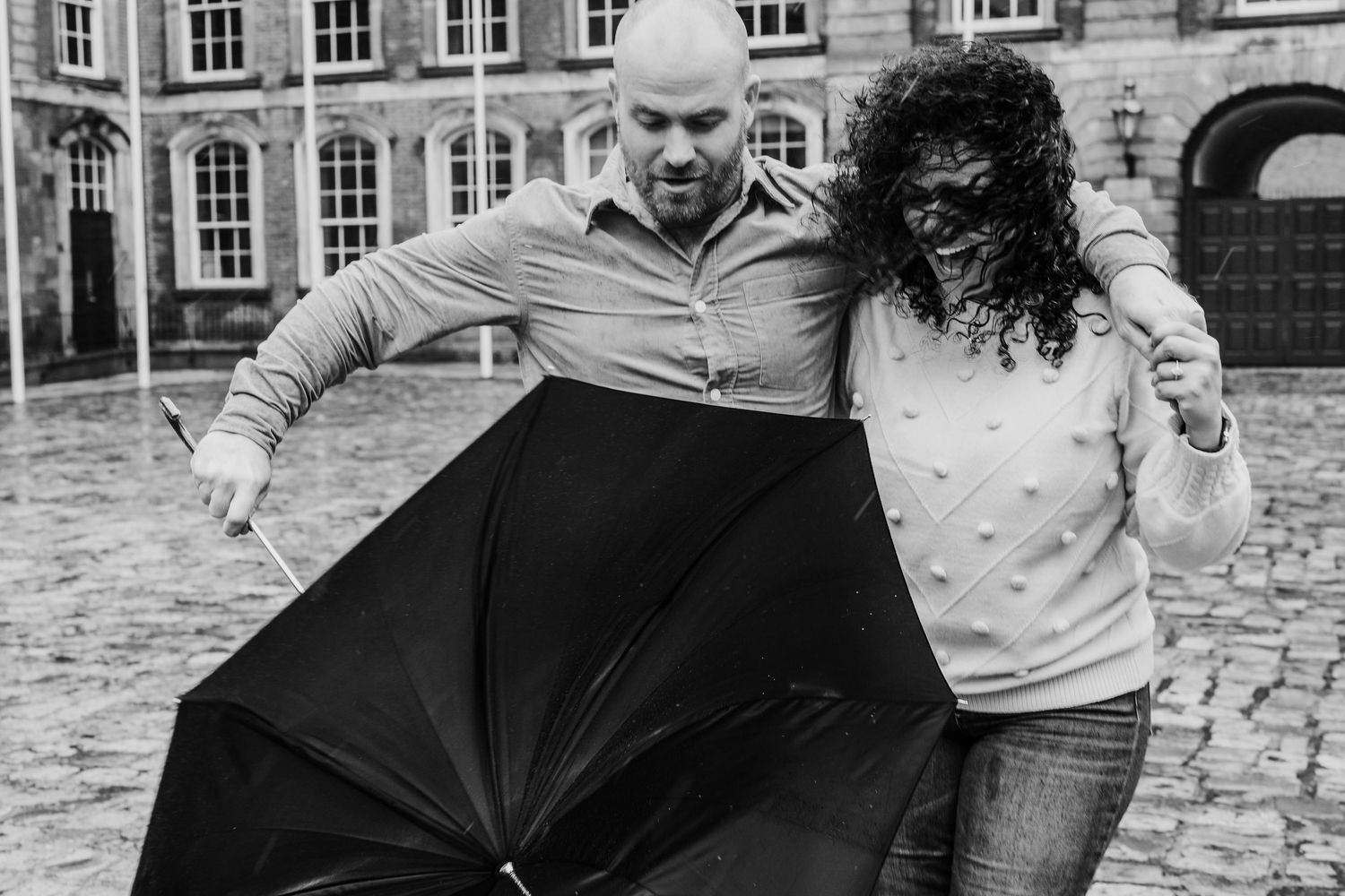 A playful couple battling with a black umbrella on a cobblestone courtyard in front of historic architecture. PHOTOGRAPHER Dublin ireland