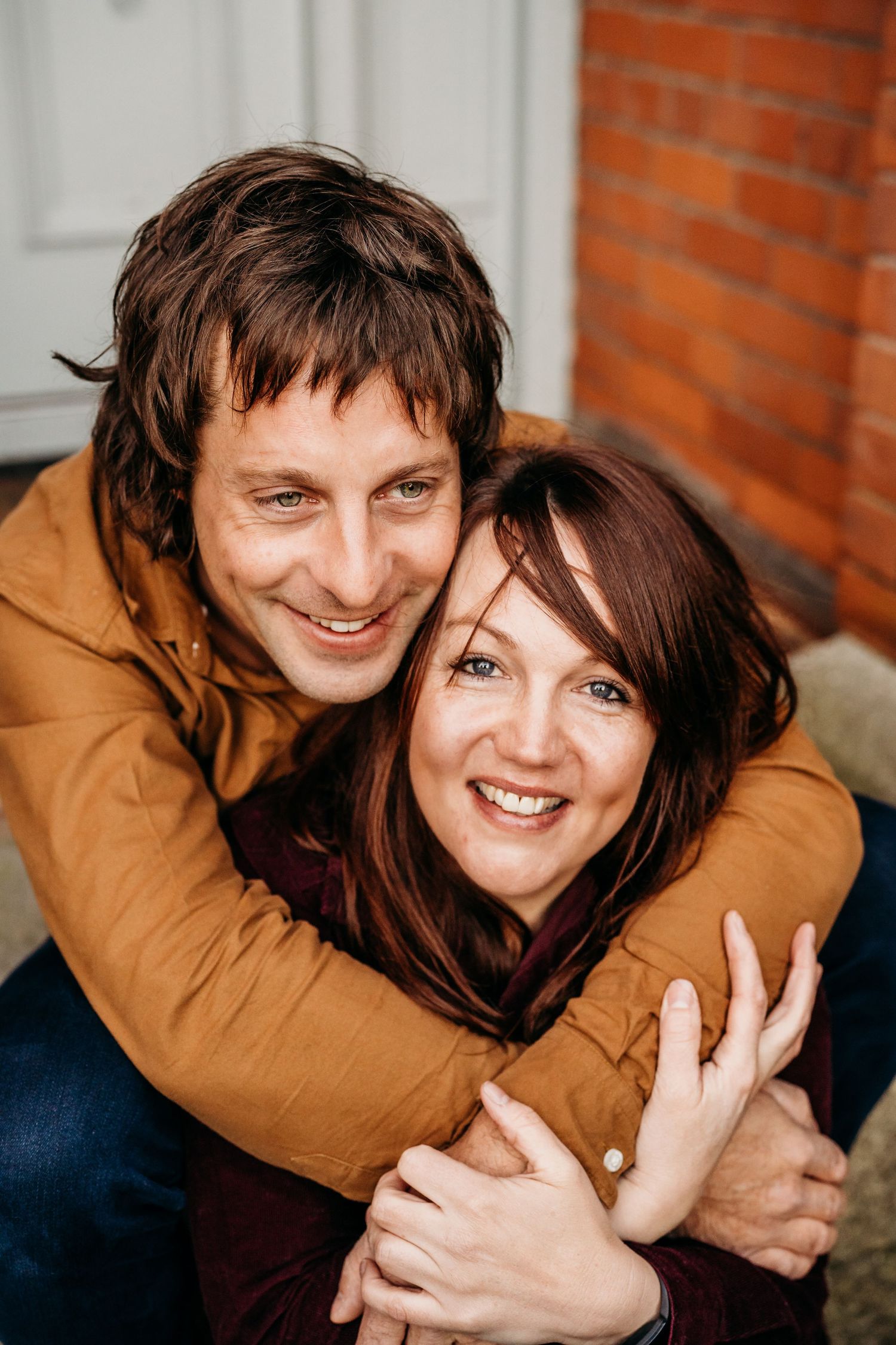 A couple embraces and smiles warmly while wearing brown and burgundy sweaters against a brick wall background. PHOTOGRAPHER Dublin ireland