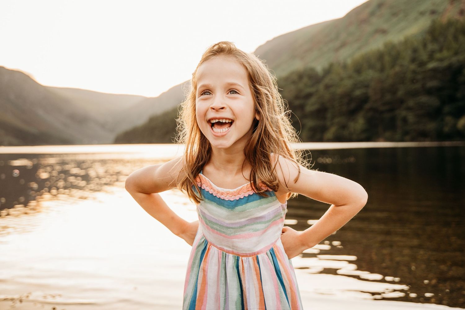 Young child laughs joyfully while playing outdoors at sunset near a tranquil lake. family PHOTOGRAPHER Dublin