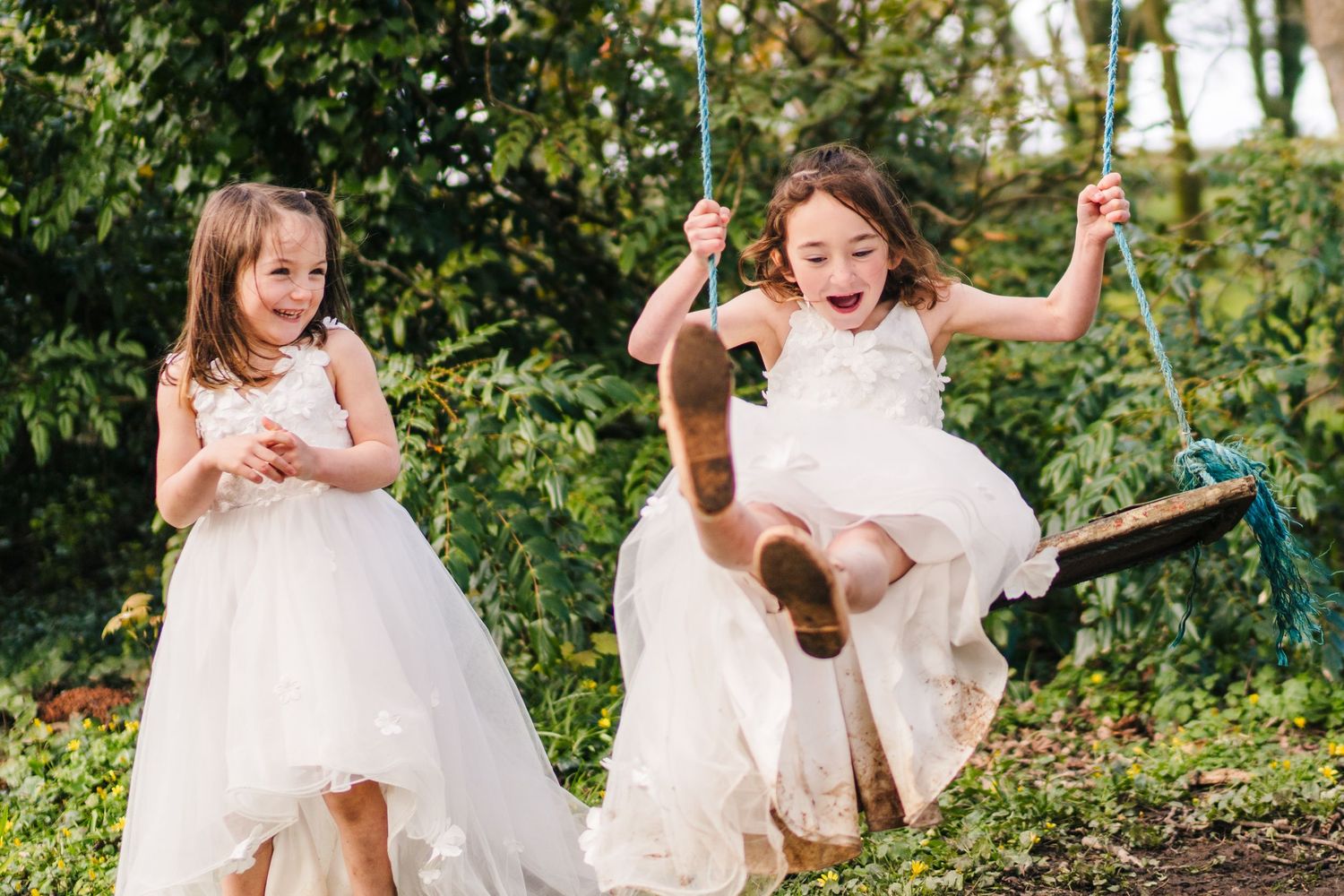 Two children in white dresses playing outdoors, with one joyfully swinging while the other stands nearby. Family PHOTOGRAPHER Dublin