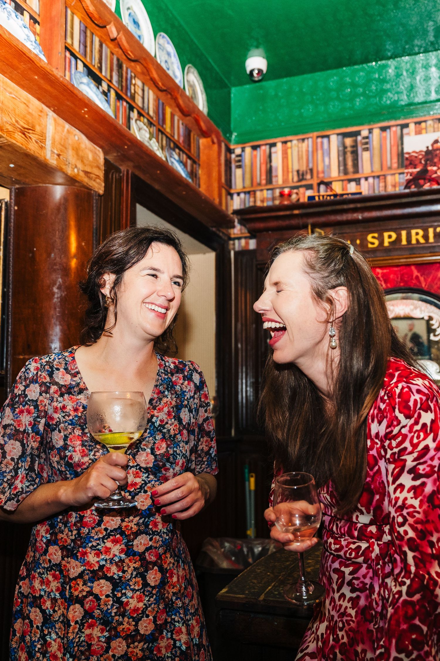 Two friends in floral dresses share drinks and laughter at a cozy pub with wooden decor and warm lighting. Wedding PHOTOGRAPHER Dublin