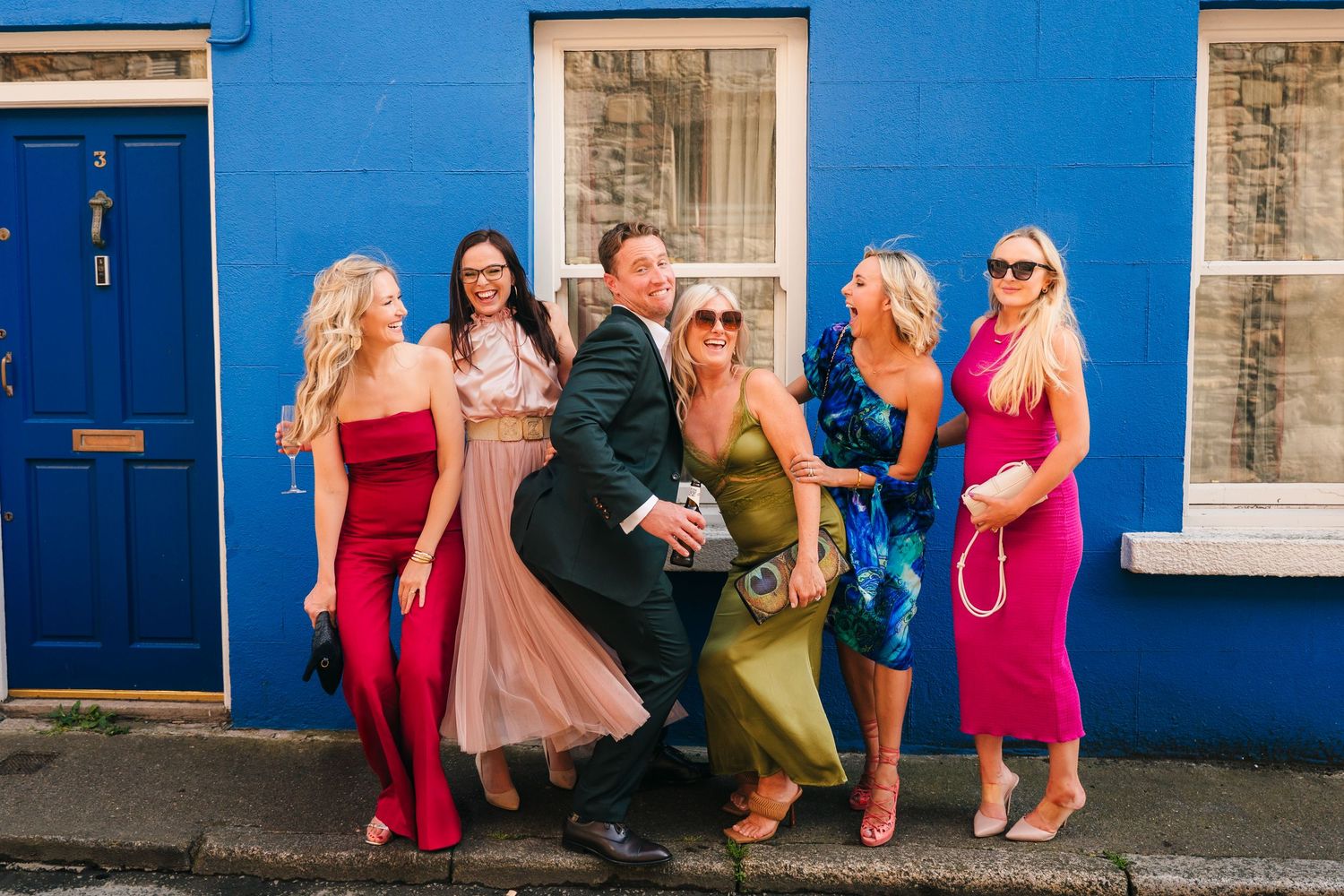 A group stands posing together against a bright blue wall with white-framed windows wearing colorful formal attire. Wedding PHOTOGRAPHER Dub