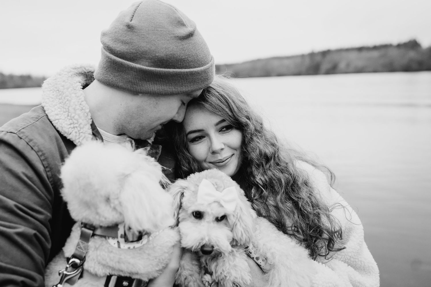 A couple snuggles close with their fluffy white dog outdoors in a black and white photograph by the water. PHOTOGRAPHER Dublin ireland