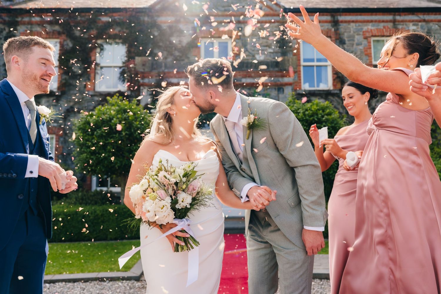Bride and groom kissing while guests throw confetti at an outdoor wedding celebration. Wedding photographer Dublin