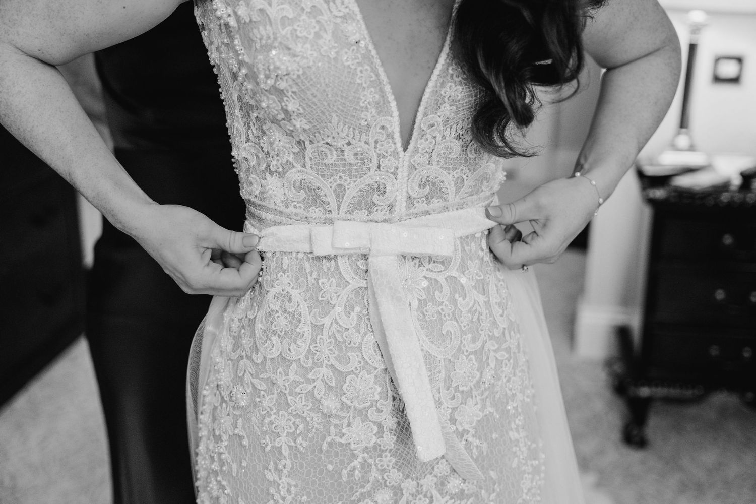 Black and white close-up of hands adjusting a delicate lace wedding dress with satin ribbon.