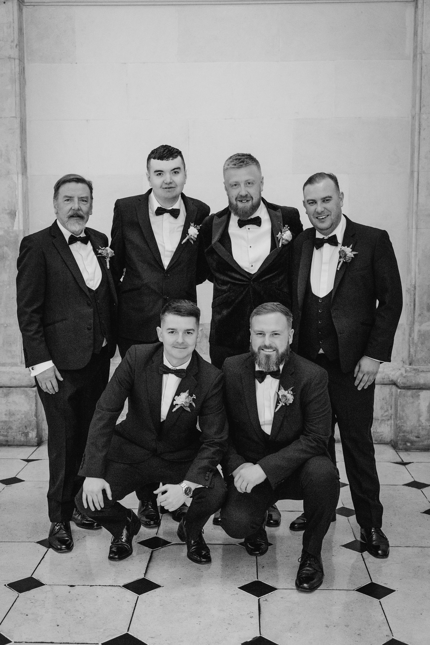 A group wearing formal black tuxedos poses together on a tiled floor at a wedding event. Wedding photographer dublin