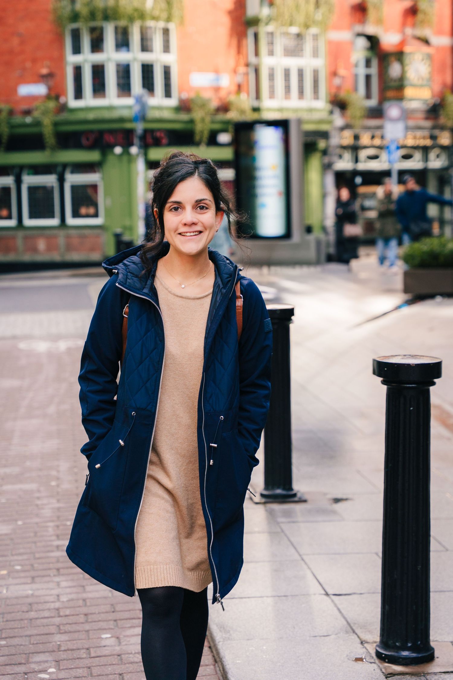 A person wearing a navy blue jacket and beige sweater poses on a brick paved street with bollards in the background. professional headshots