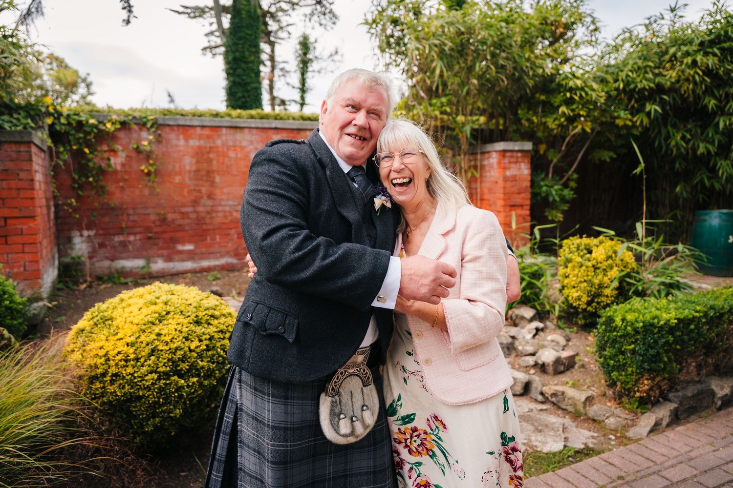 Couple sharing a happy moment in garden with brick wall and Scottish kilt attire at wedding celebration. Portmarnock Hotel Wedding PHOTOGRAP