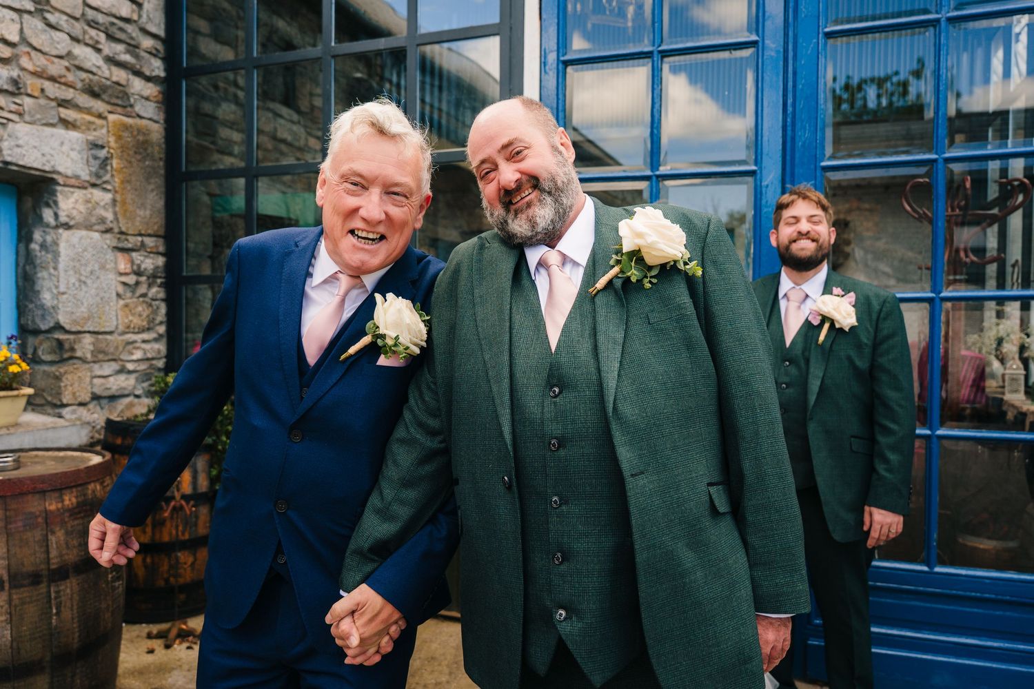 People laugh and smile while wearing formal suits outside a rustic building with blue doors and stone walls. . Village Barn Tyrrellspass wed