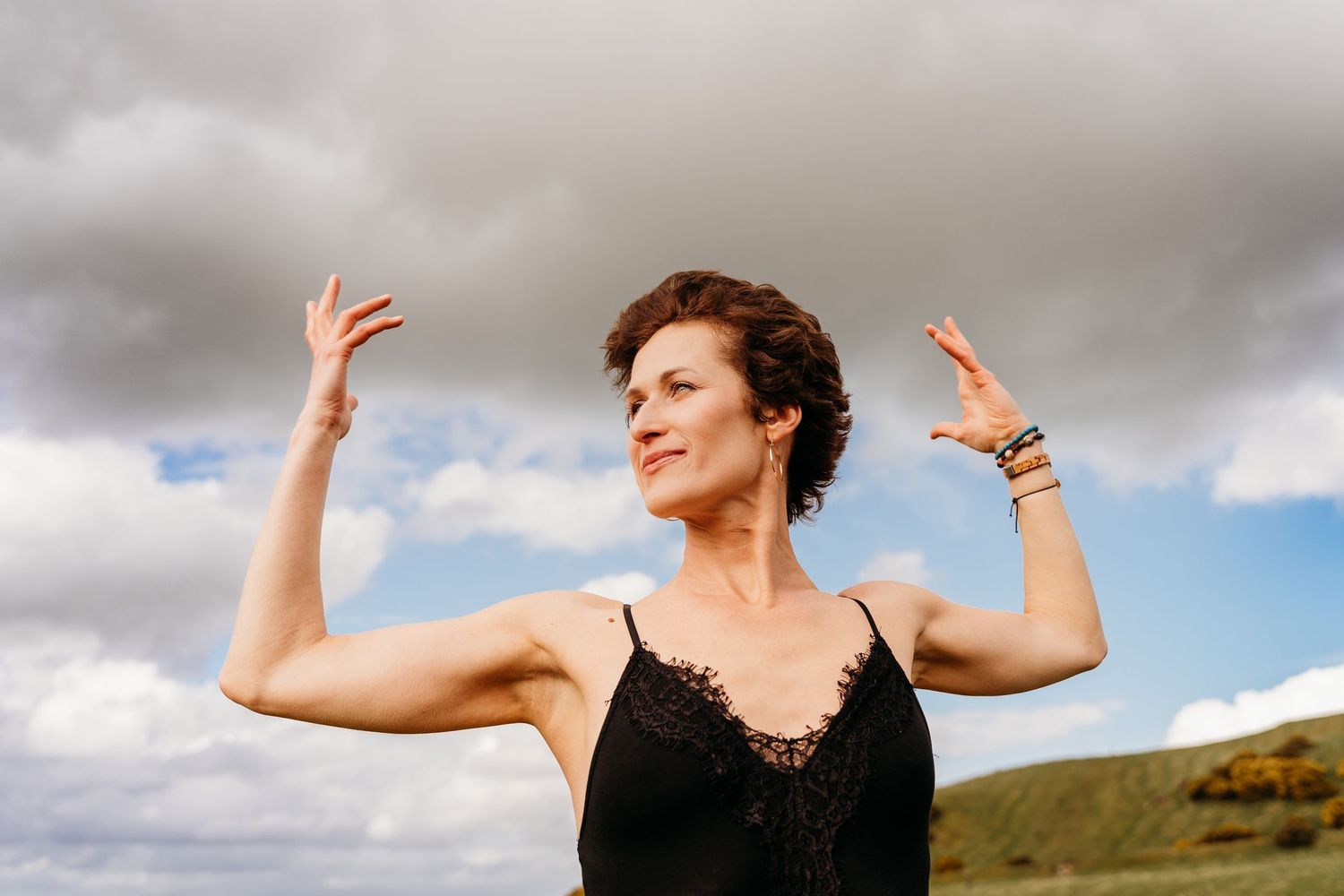 Person in black dress raises arms against dramatic cloudy sky and green hillside landscape. Portrait PHOTOGRAPHER Dublin