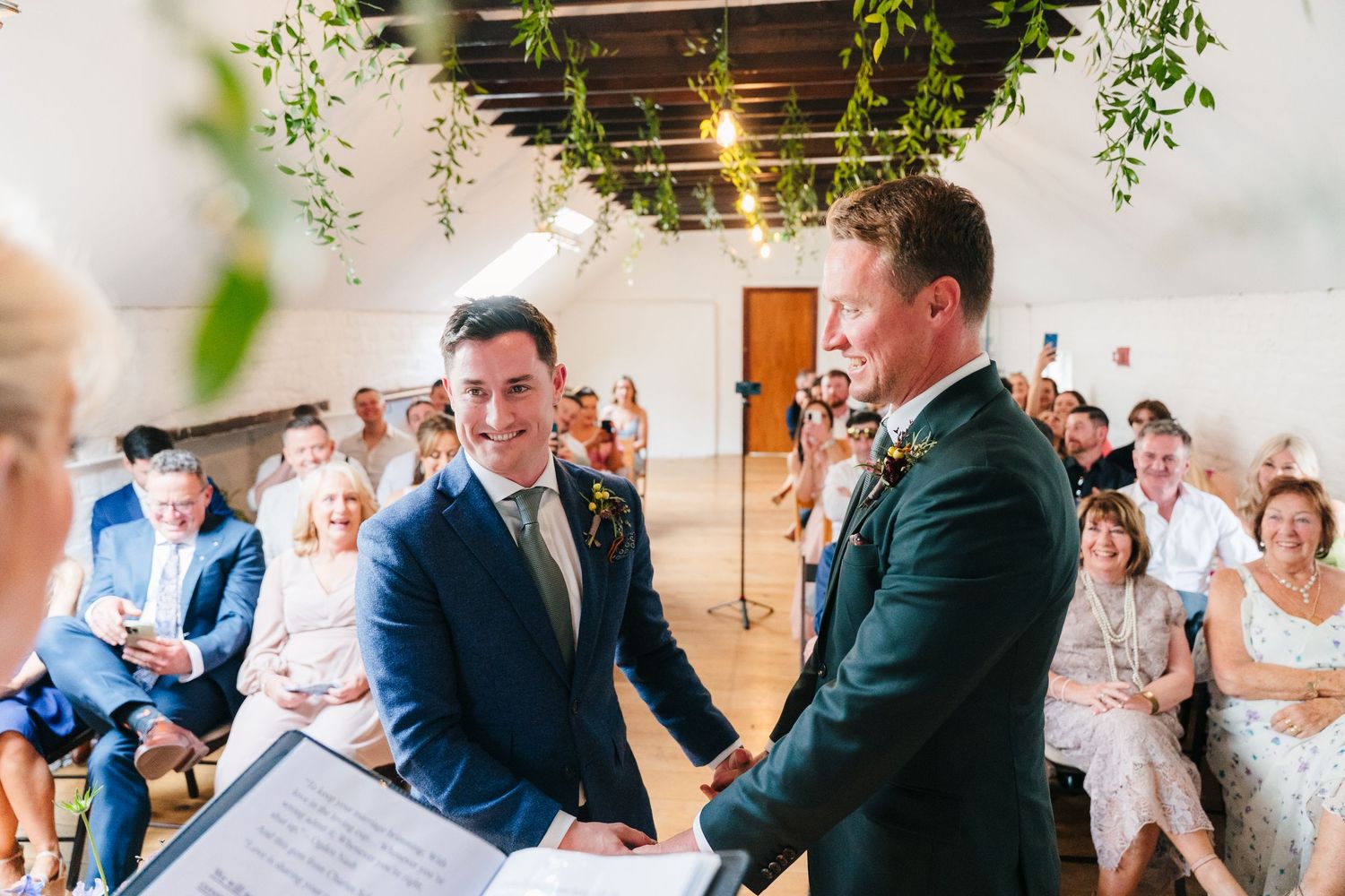 Wedding party celebrating in a bright indoor venue with hanging greenery. Same sex wedding PHOTOGRAPHER Dublin