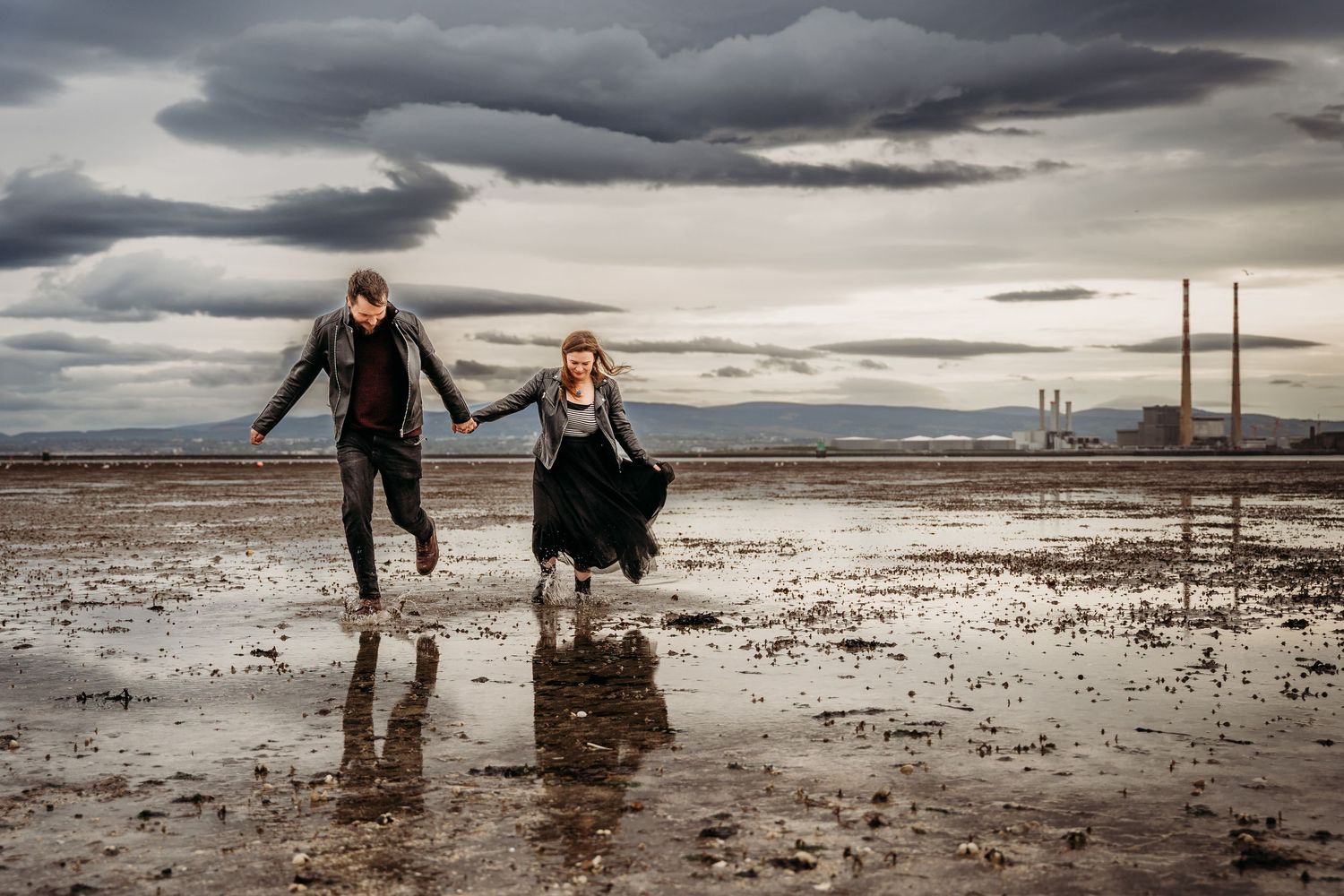 People play and splash in shallow coastal waters with industrial chimneys visible on the horizon. engagement PHOTOGRAPHER Dublin