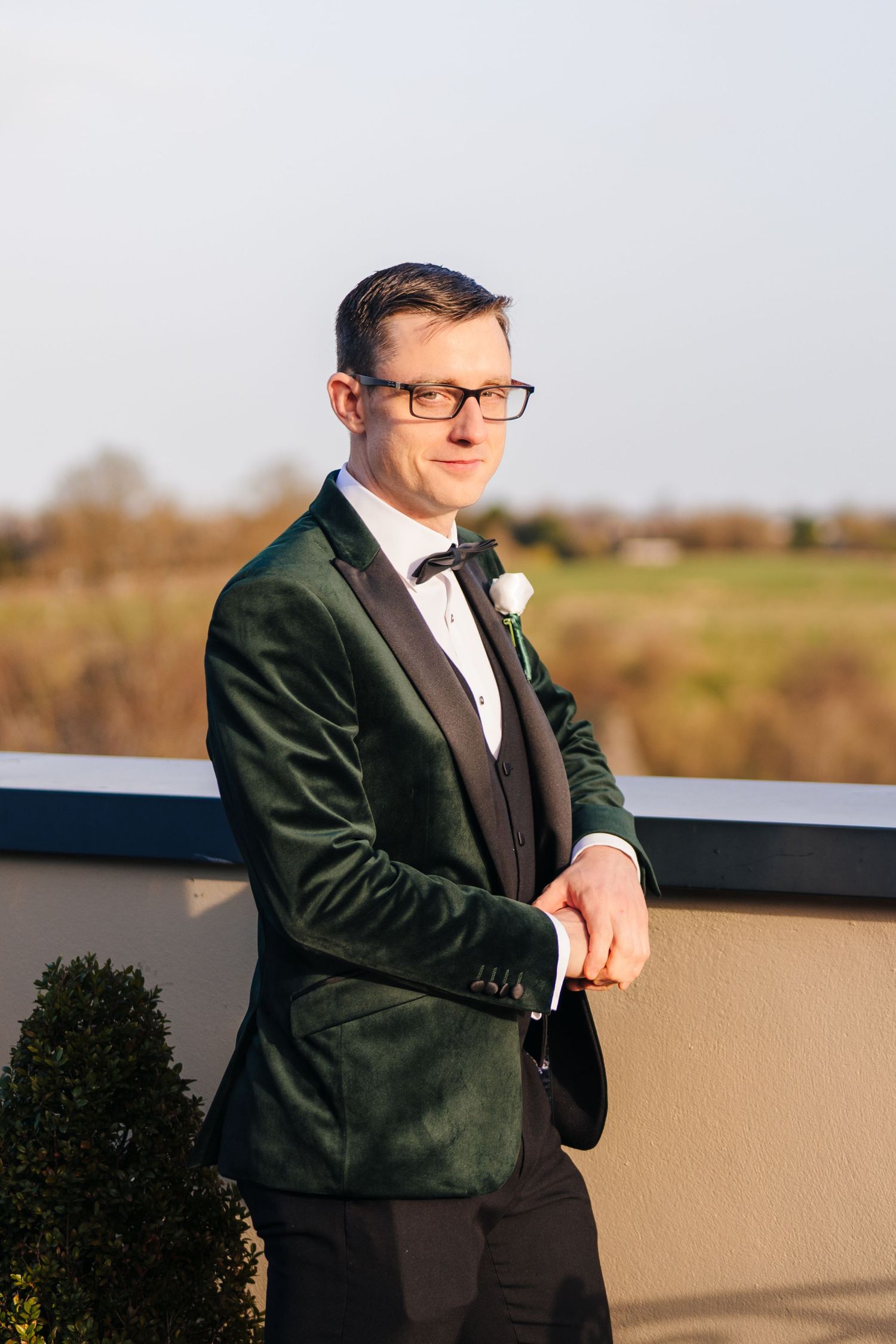 A groom in a dark green velvet tuxedo jacket and bow tie poses outside during golden hour.  Wedding PHOTOGRAPHER Trim Castle