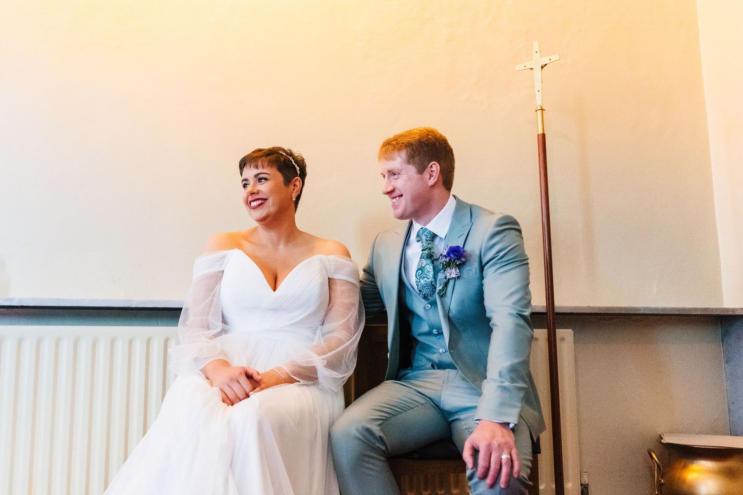 A tender wedding moment shows a couple sitting together indoors, with the bride in white and groom in a light gray suit.Millhouse slane wedd