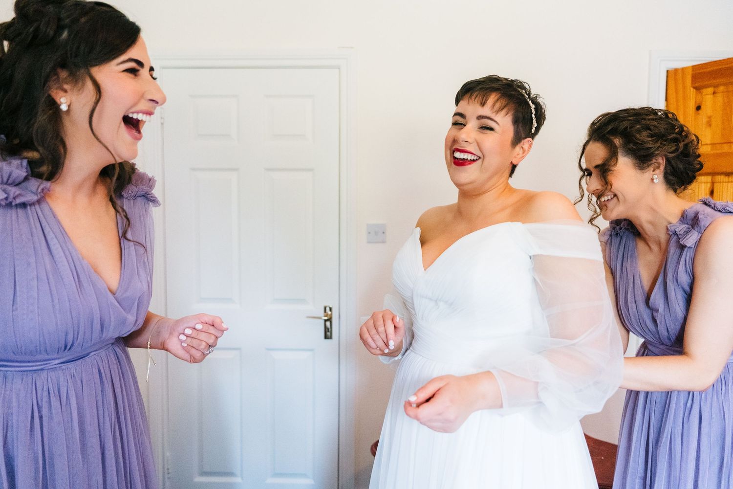 Three figures in purple and white dresses share a heartfelt laugh during a special occasion. Millhouse slane wedding photographer dublin