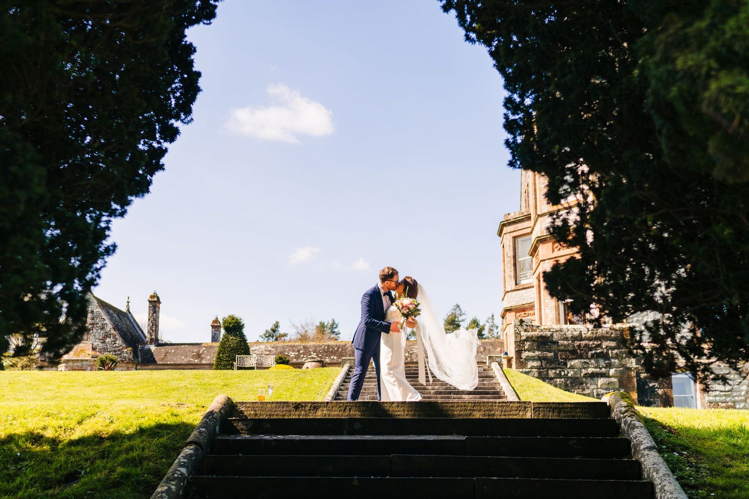 Couple sitting on grand stone steps of a cathedral with a scenic countryside view on a sunny day. Castle Leslie Wedding Photographer Dublin