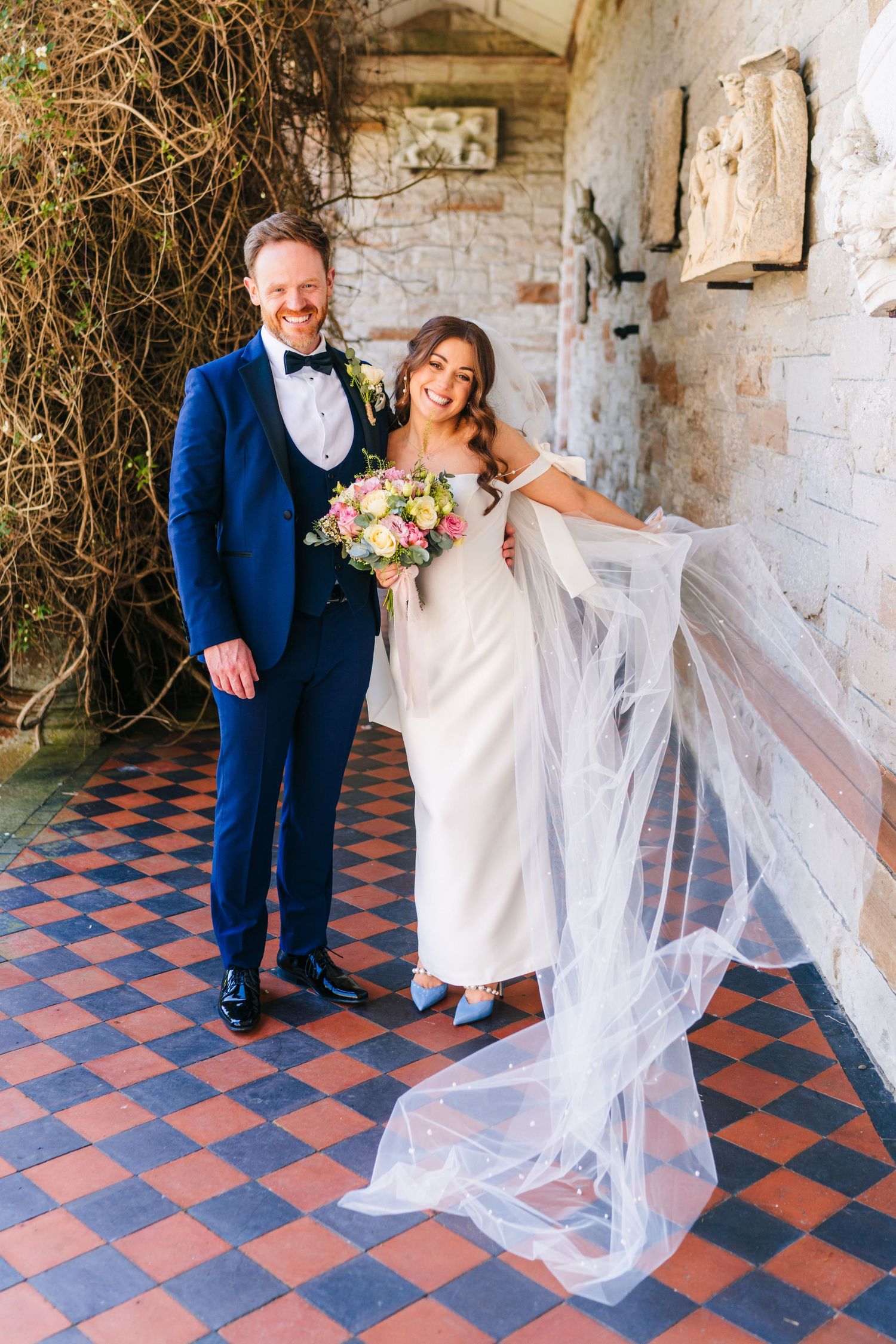 Castle Leslie A newlywed couple poses in a rustic hallway with tiled floors and exposed brick walls as the bride's veil flows dramatically.