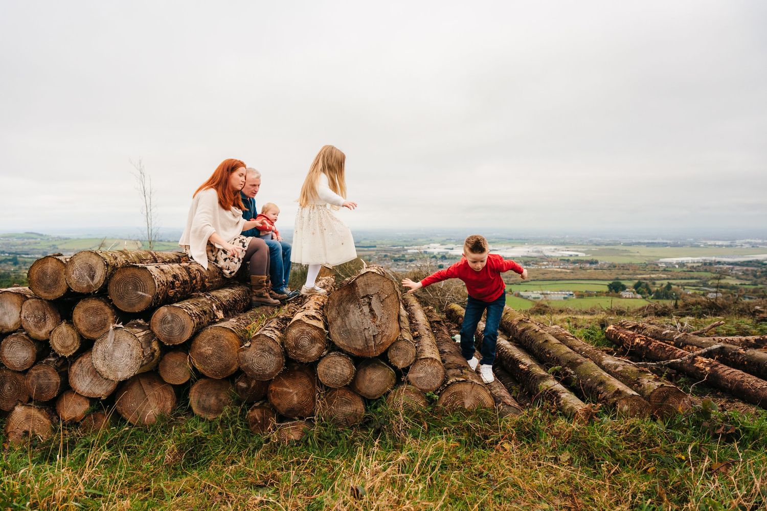 Family Photographer Dublin Ireland A group sits together on stacked logs overlooking a scenic countryside vista.