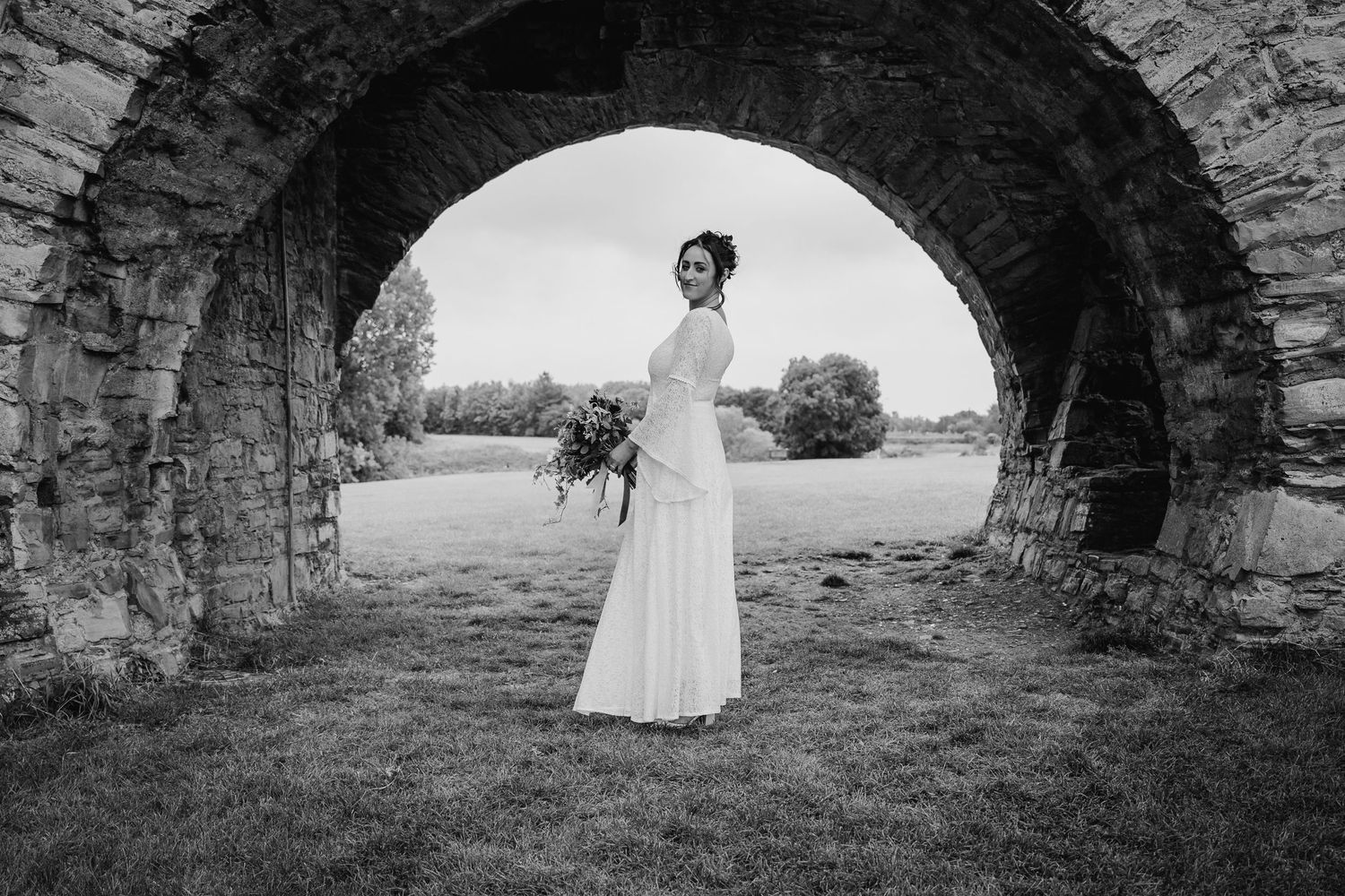 Bride posing in flowing white dress under historic stone archways in black and white photography. v