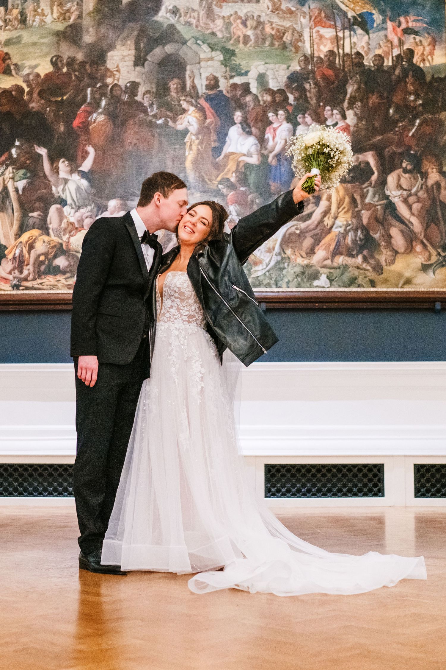 A bride in a leather jacket poses with their partner in front of a dramatic Renaissance-style mural. Wedding PHOTOGRAPHER Dublin
