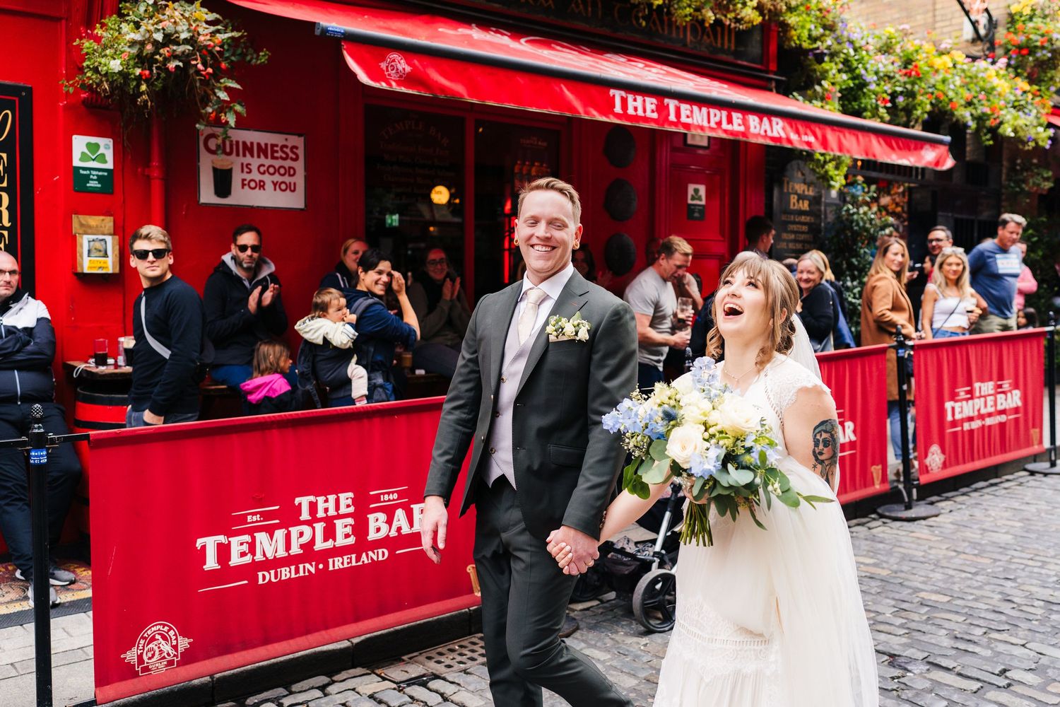 Newlyweds walking past red Temple Bar pub in Dublin with floral decorations. PHOTOGRAPHER Dublin Ireland Wedding