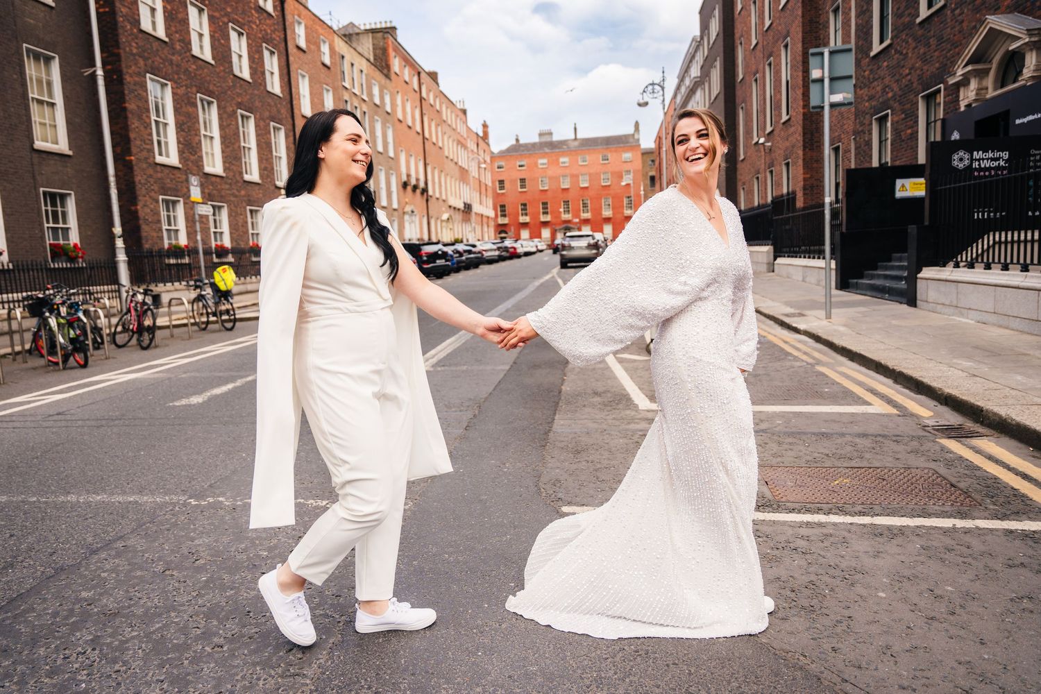 A couple in white outfits holding hands playfully on a Dublin street with classic brick buildings in the background. House Dublin Wedding Ph