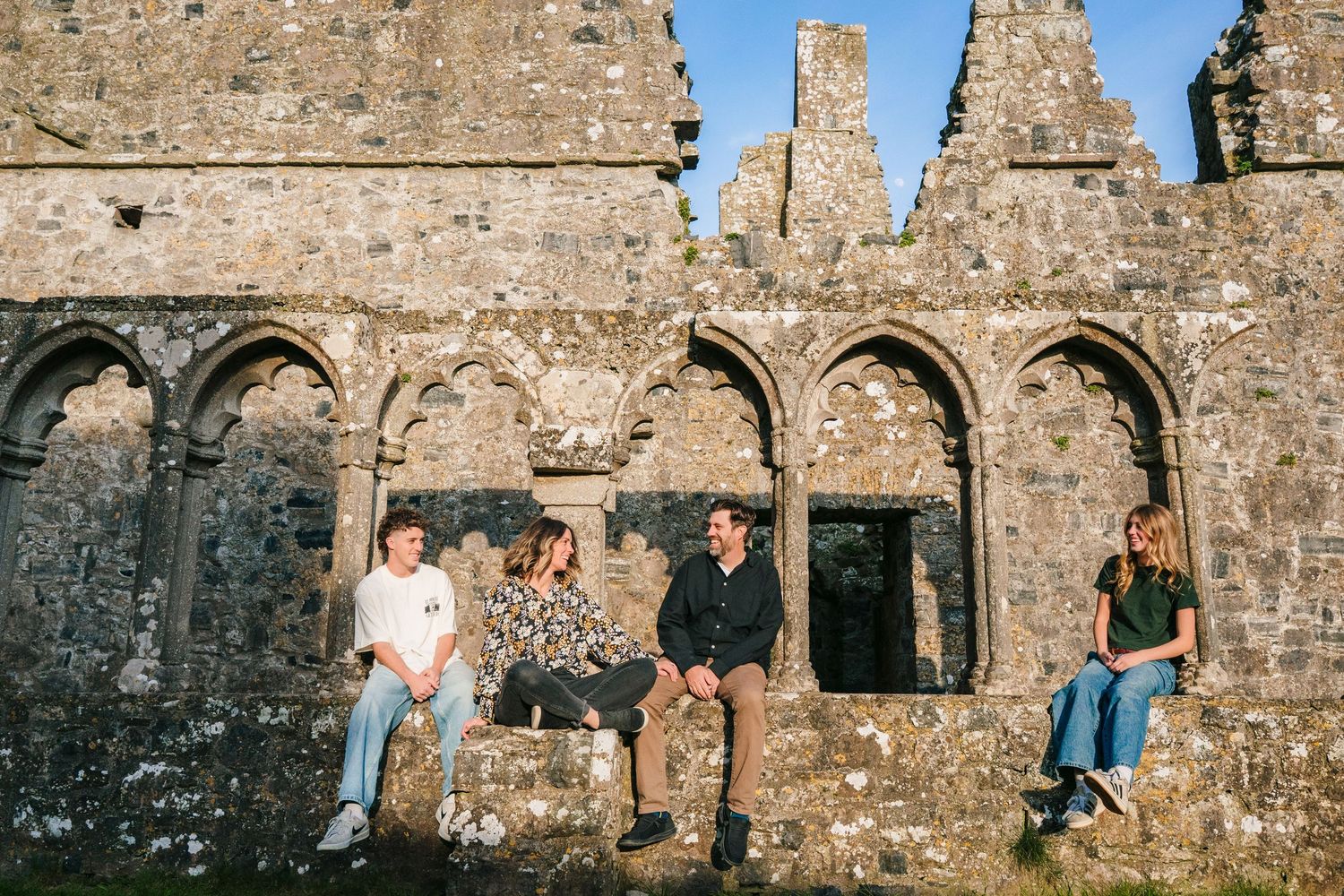 A group relaxes along a stone wall of ancient ruins with gothic arched windows and weathered stone architecture. Photographer Ireland dublin