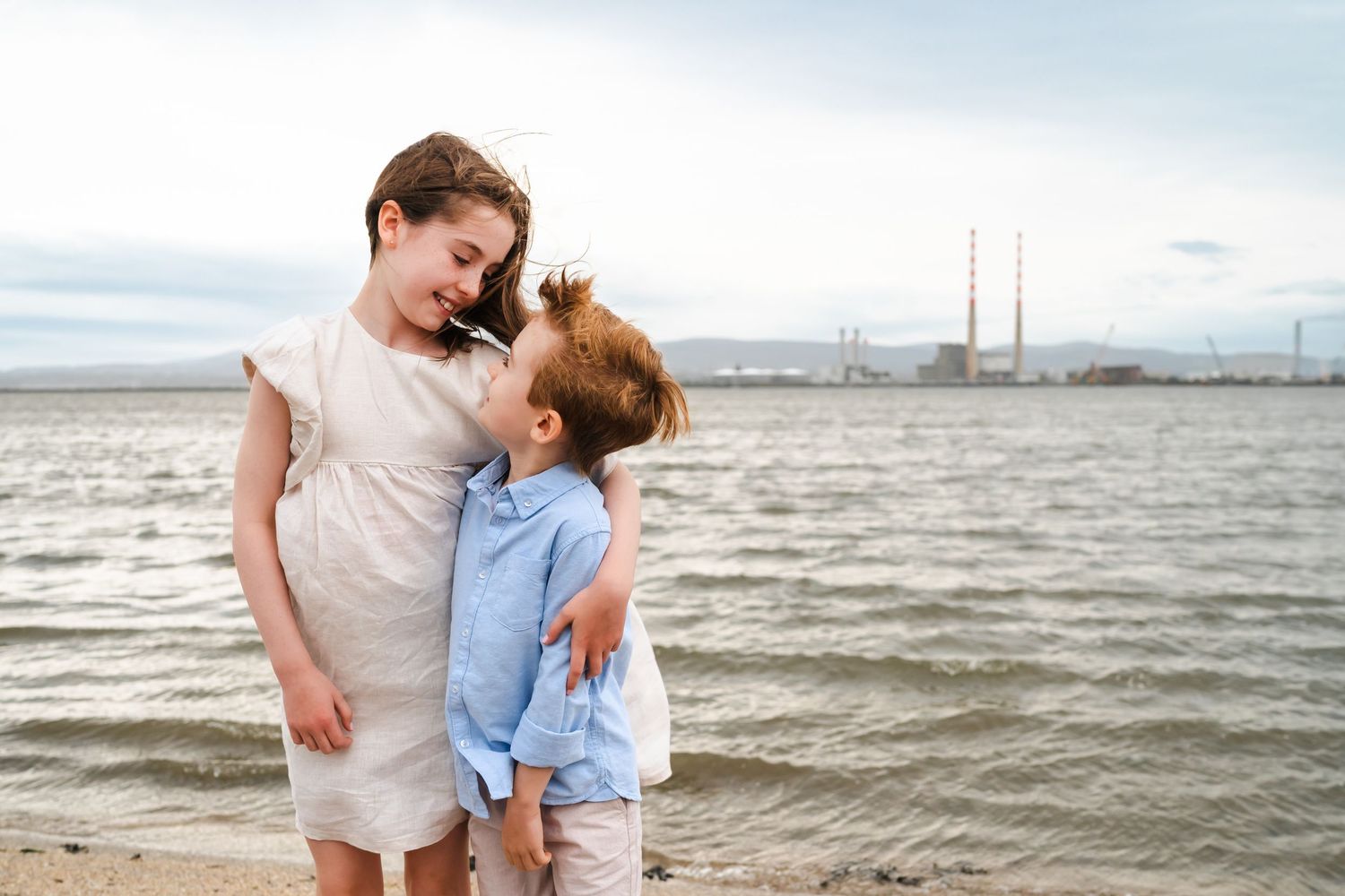 A parent and child wearing casual clothing share a tender moment while walking along a scenic beach at sunset.  Family photographer Dublin I