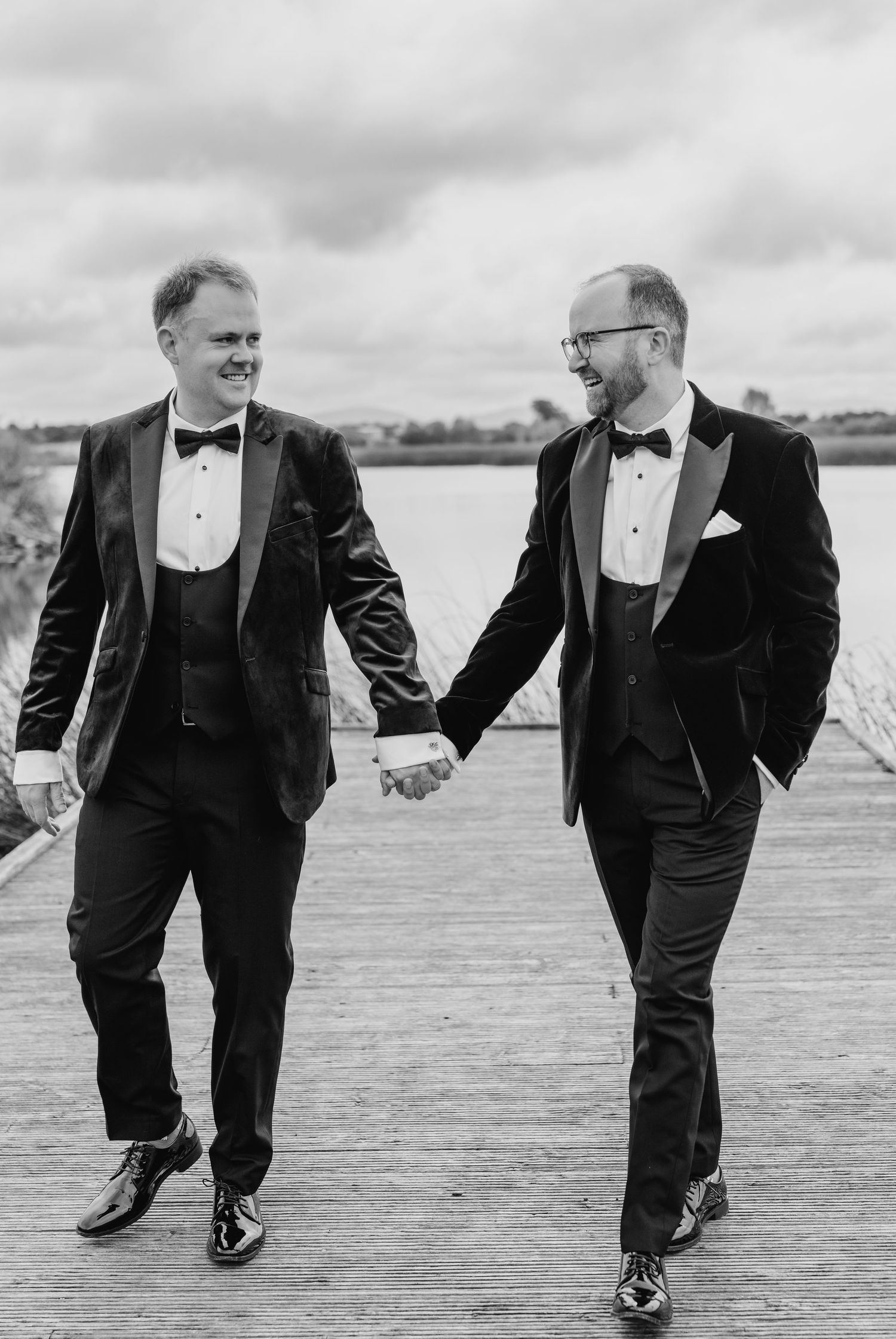 Two people in tuxedos walking and holding hands on a wooden boardwalk by the water. LGBTQ Ashley Park Wedding Photographer
