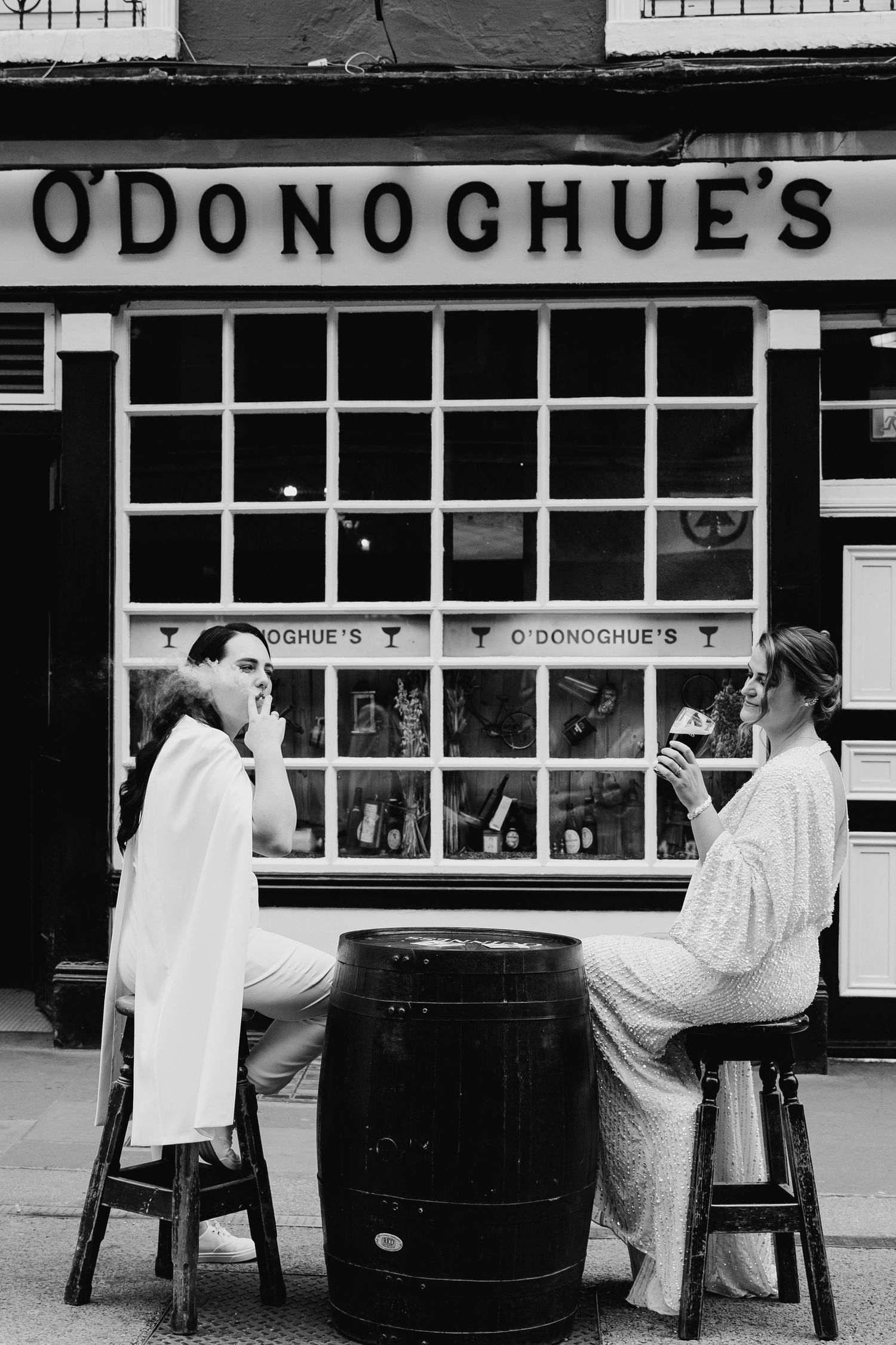 Two people in white clothing sit at a barrel outside O'Donoghue's pub storefront in black and white photograph. Wedding PHOTOGRAPHER Dublin