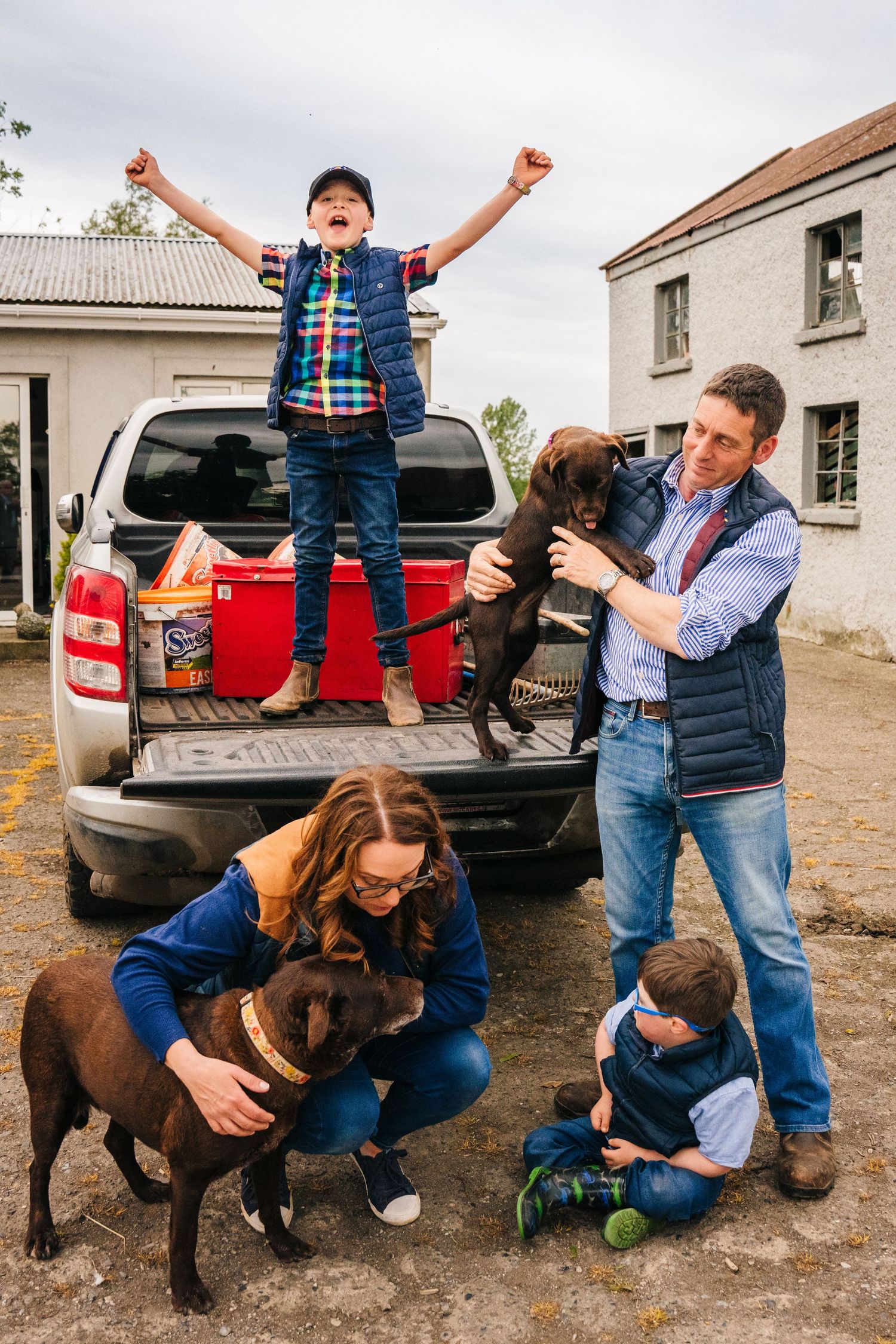 Family enjoying time outdoors with their chocolate labrador dogs around pickup truck bed on farmyard property. family PHOTOGRAPHER Dublin