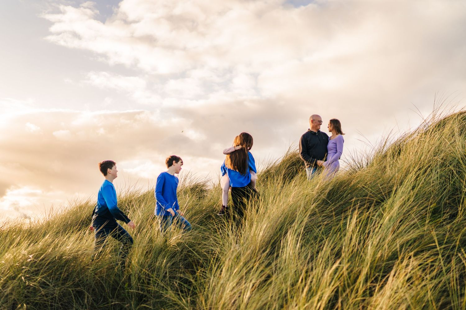 People hiking through tall wind-swept grass on a coastal hillside during golden hour with dramatic clouds overhead. Family PHOTOGRAPHER Dubl