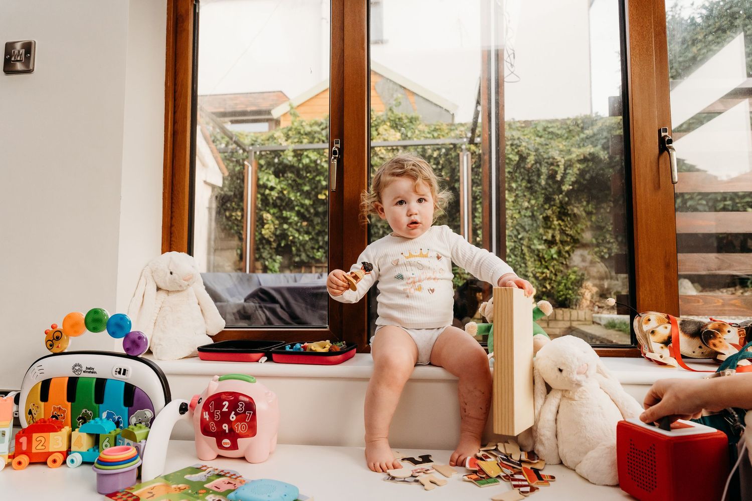 Toddler plays with toys near wooden windows in living room surrounded by stuffed animals and colorful toys. family PHOTOGRAPHER Dublin