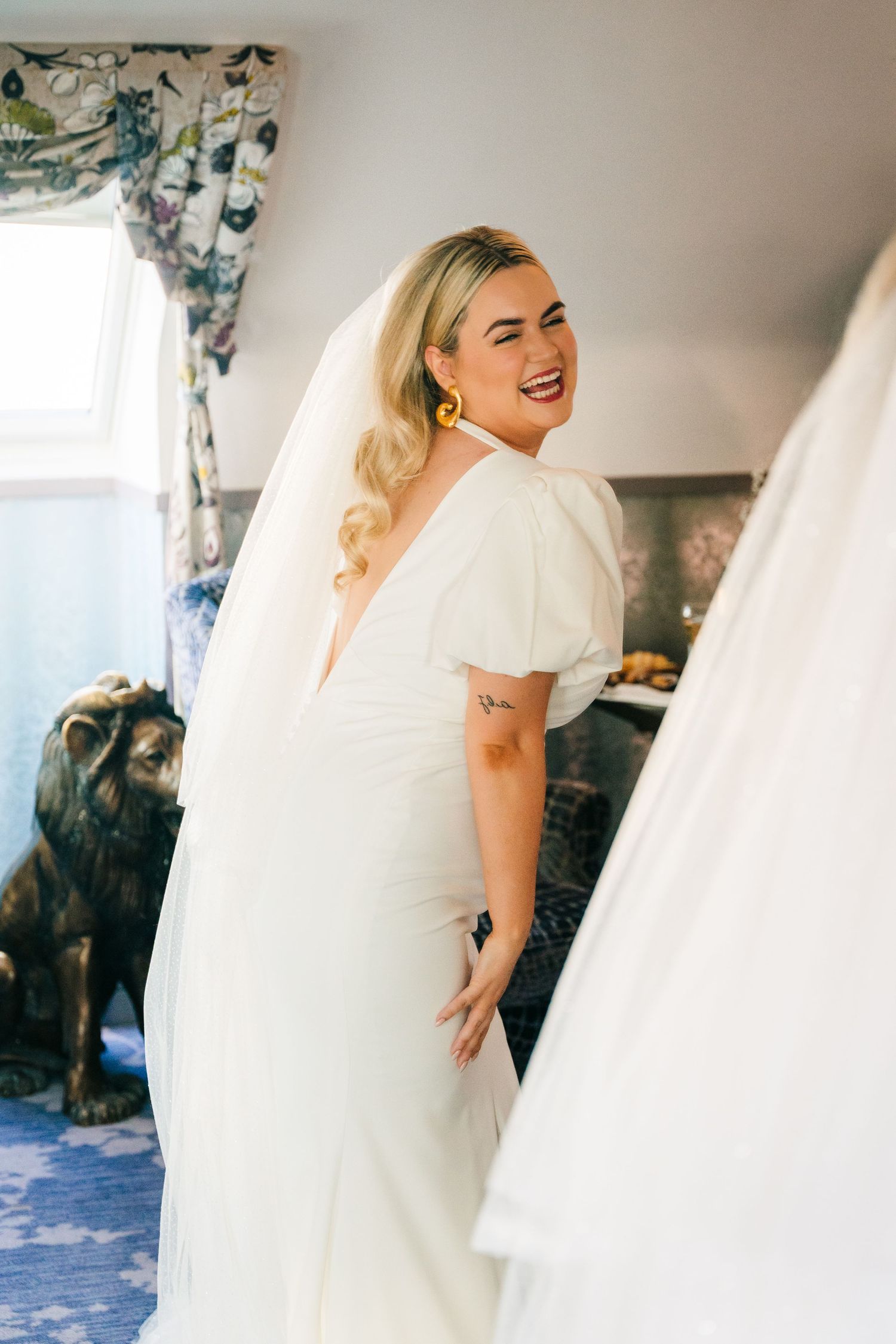 A joyful moment captured during wedding preparations as a bride in white laughs while getting ready in a room with floral curtains. Wedding