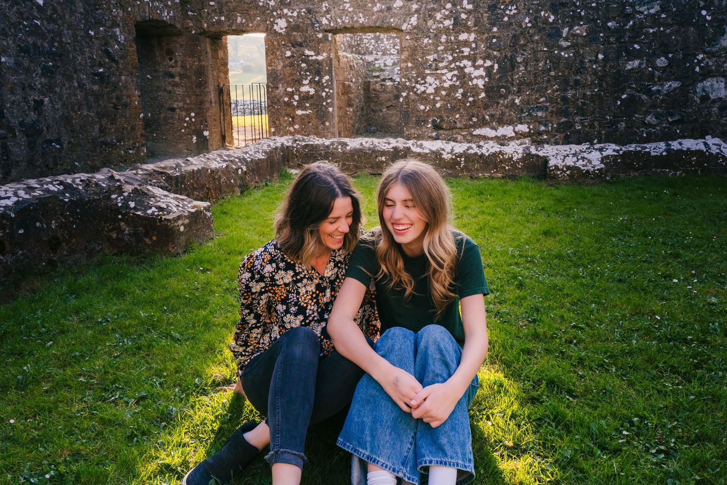 Two friends sit together on grass in front of an old stone wall, wearing denim skirts in warm sunlight. Photographer Ireland dublin