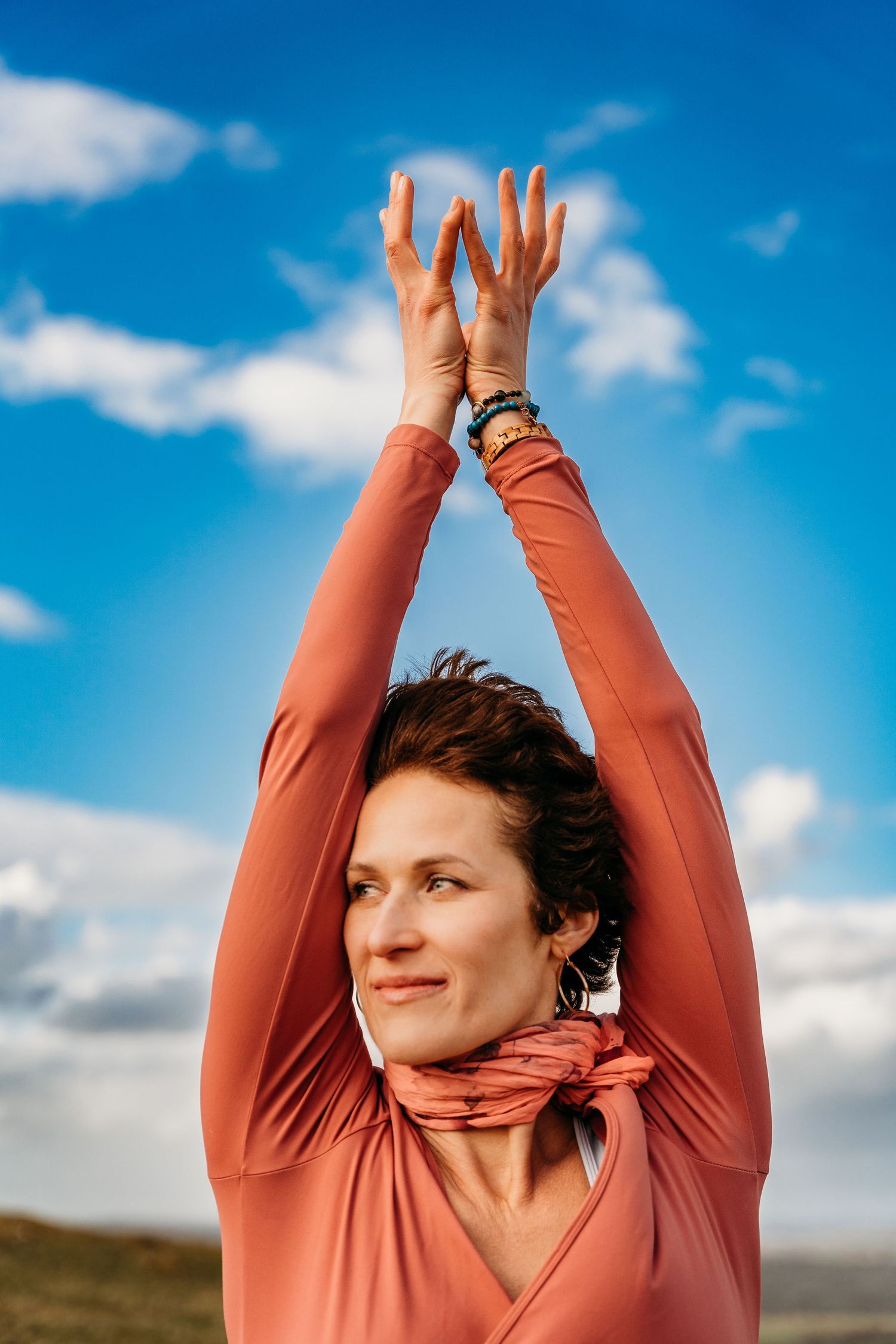 Person in coral-colored top stretches arms upward against blue sky with white clouds on sunny day. Portrait PHOTOGRAPHER Dublin