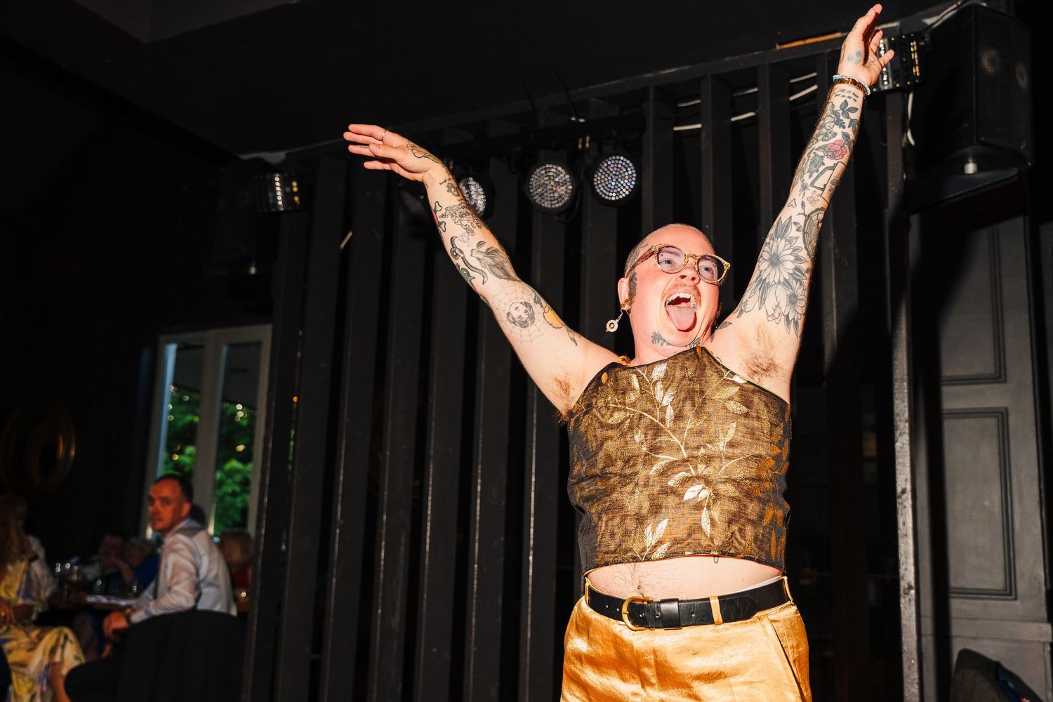 Poulaphouca Wedding photographer wicklow  Person in gold top and tattoos dances enthusiastically with arms raised at an indoor event.